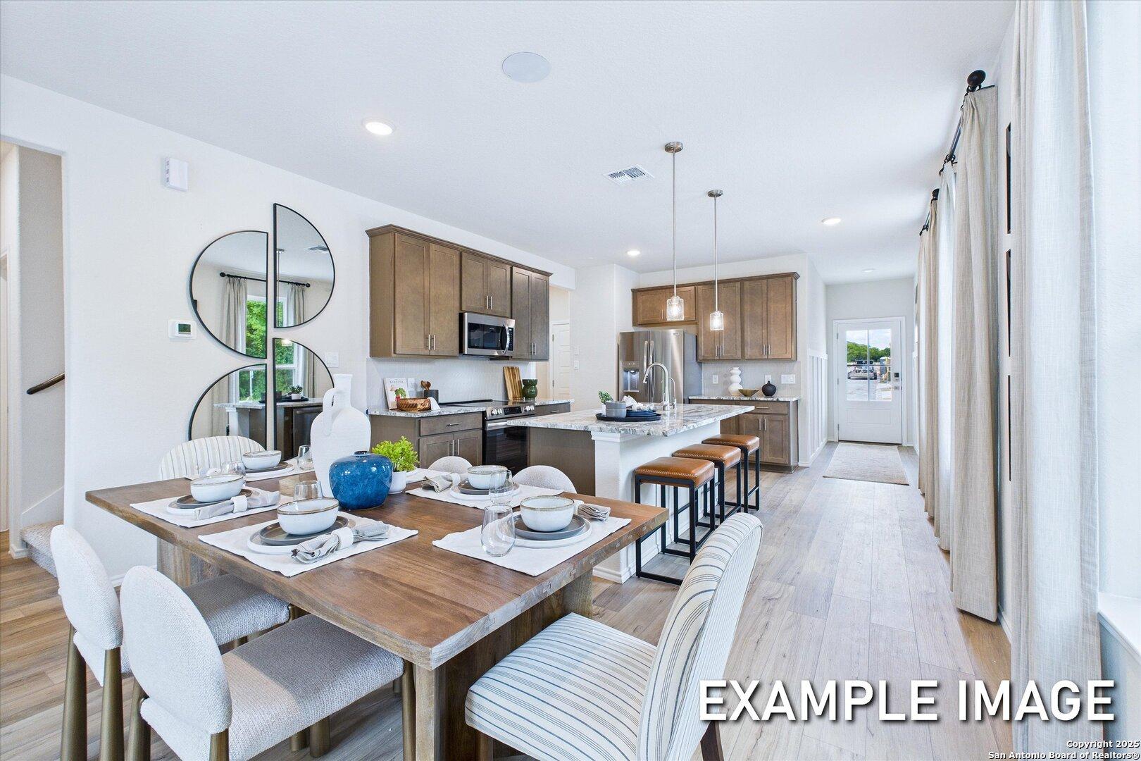Open-concept dining area with wooden table and white chairs adjacent to modern kitchen with quartz island, stainless appliances, and shaker cabinets in The Gillian C, San Antonio
