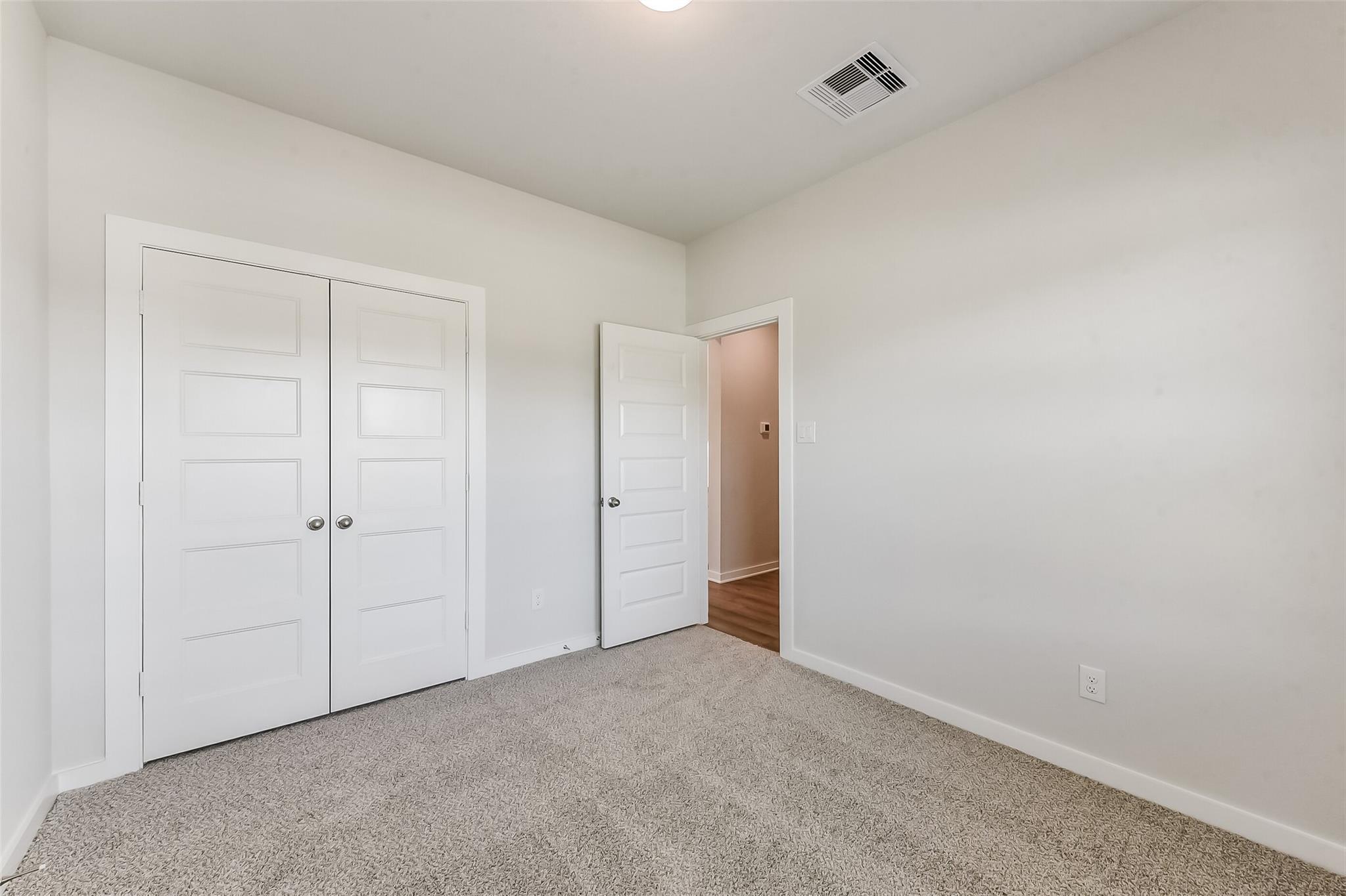 Bright bedroom featuring white walls, beige carpet, double closet doors, and open bathroom entry in Davidson Homes The Costa C, Dayton, Texas