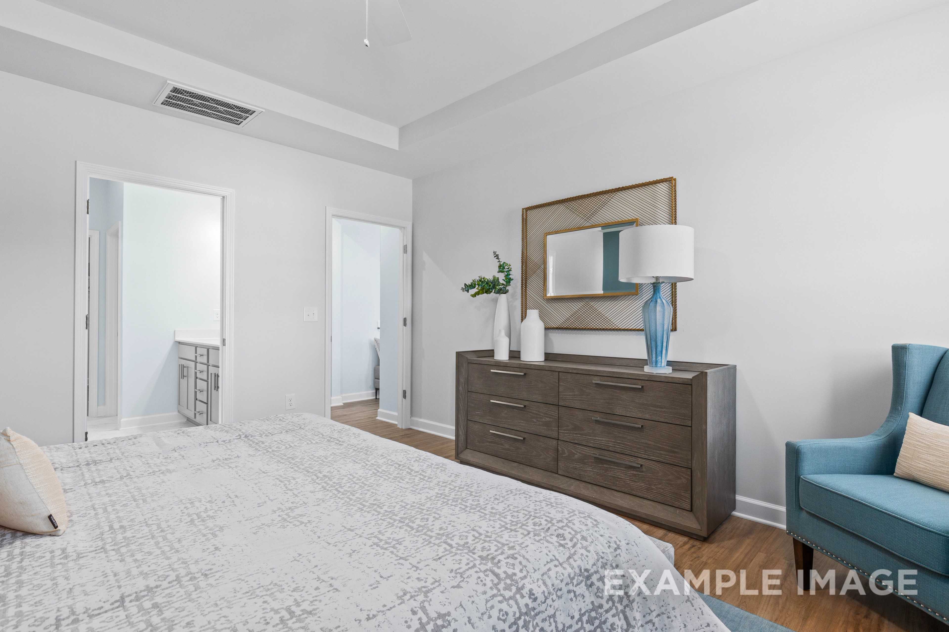 Spacious master bedroom in The Ash C with king bed, wooden dresser, blue armchair, lamp, and ensuite doorway
