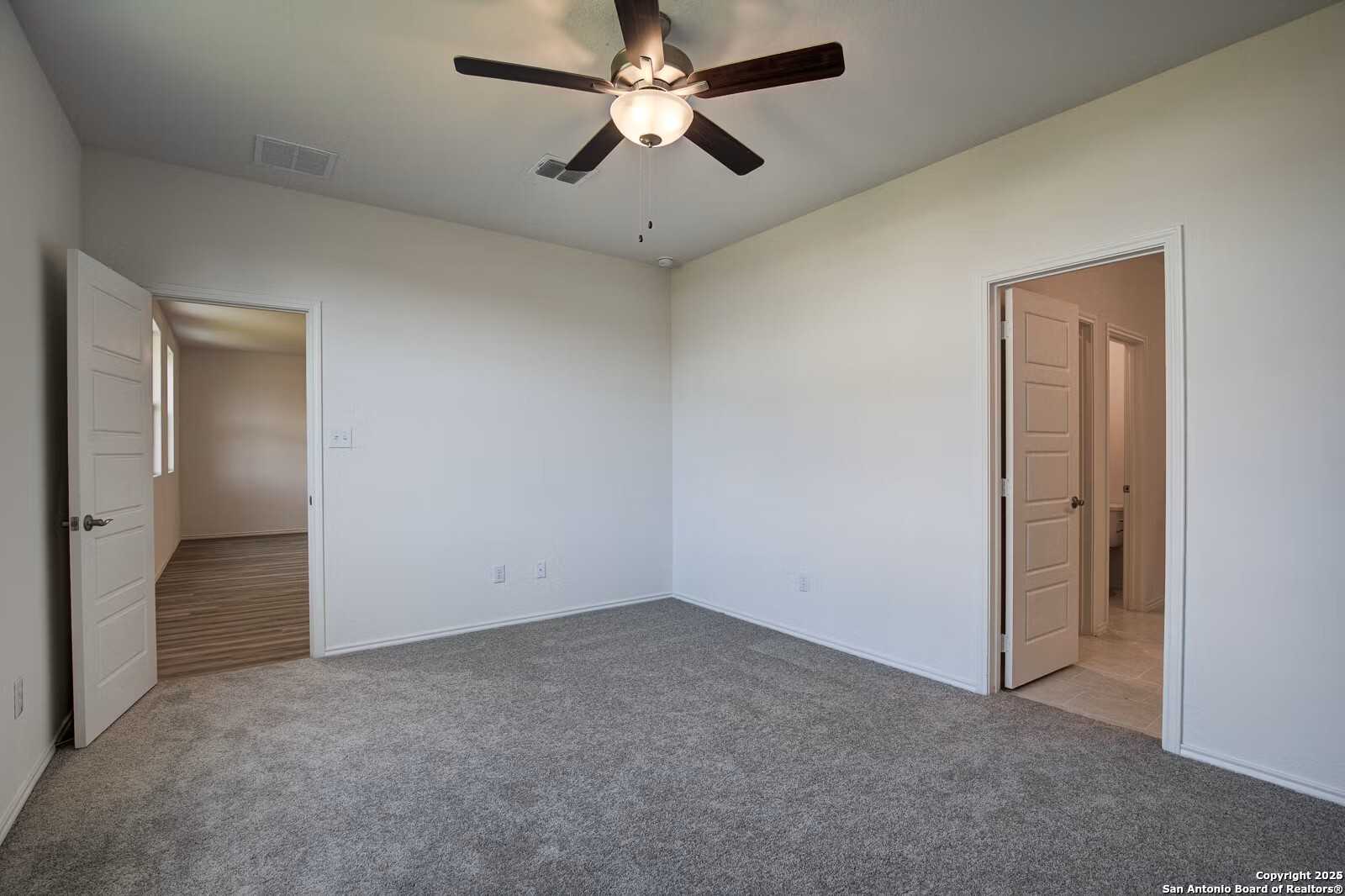 Spacious bedroom with ceiling fan, gray carpet, and adjacent bathroom in Davidson Homes Douglas B, Seguin, Texas
