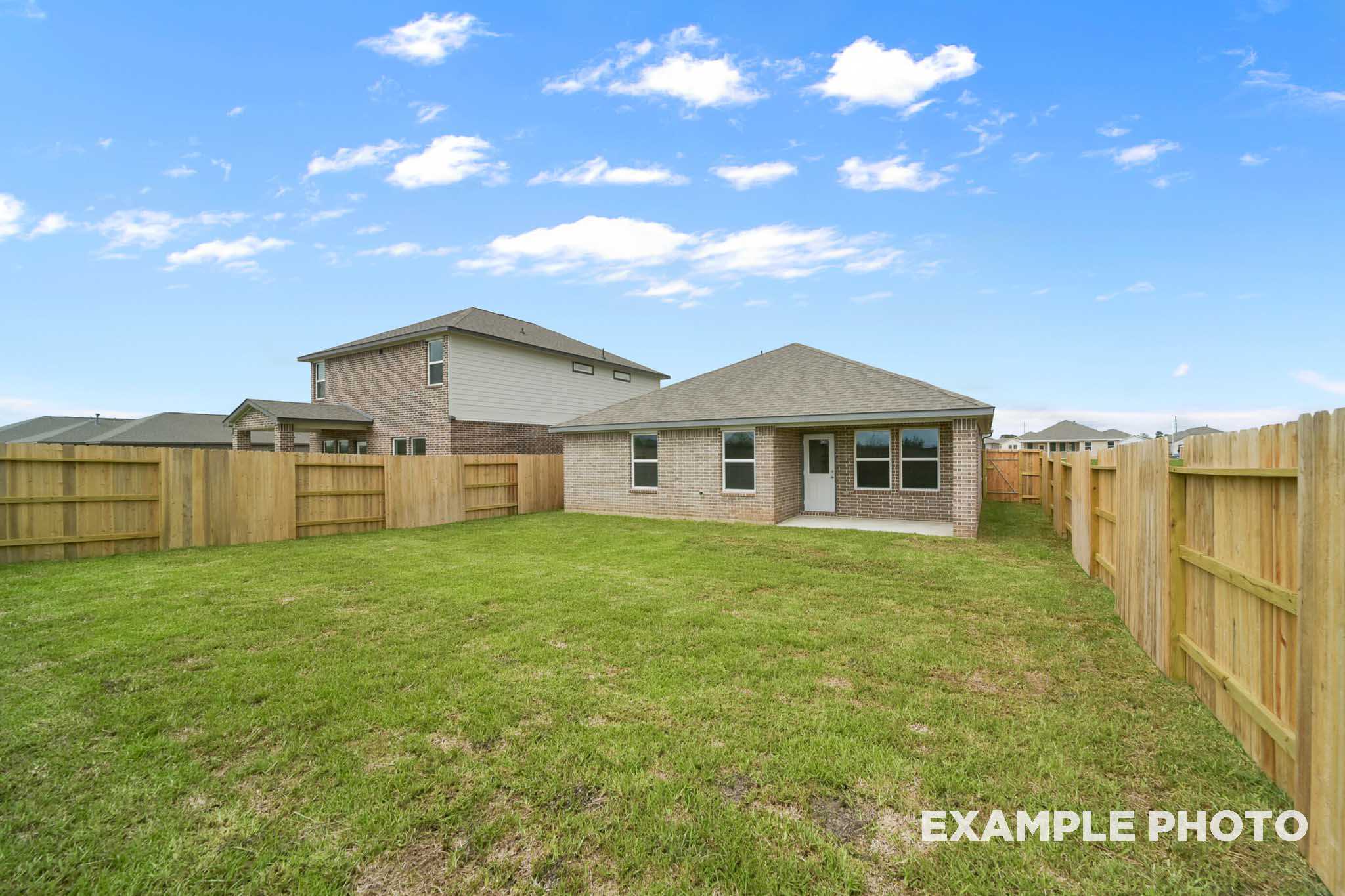 Spacious backyard of The Costa C single-story home with brick exterior, wooden fence, lush green lawn, and blue sky in Beasley, Texas