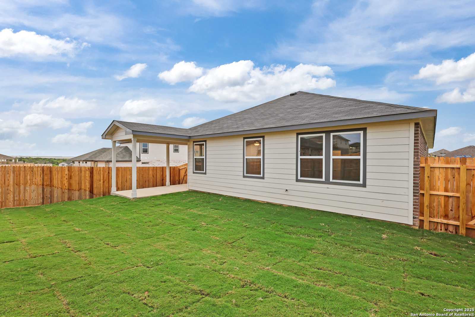 Rear view of The Daphne K single-story home with covered patio, large windows, green lawn, and wood fence in Comanche Ridge, San Antonio