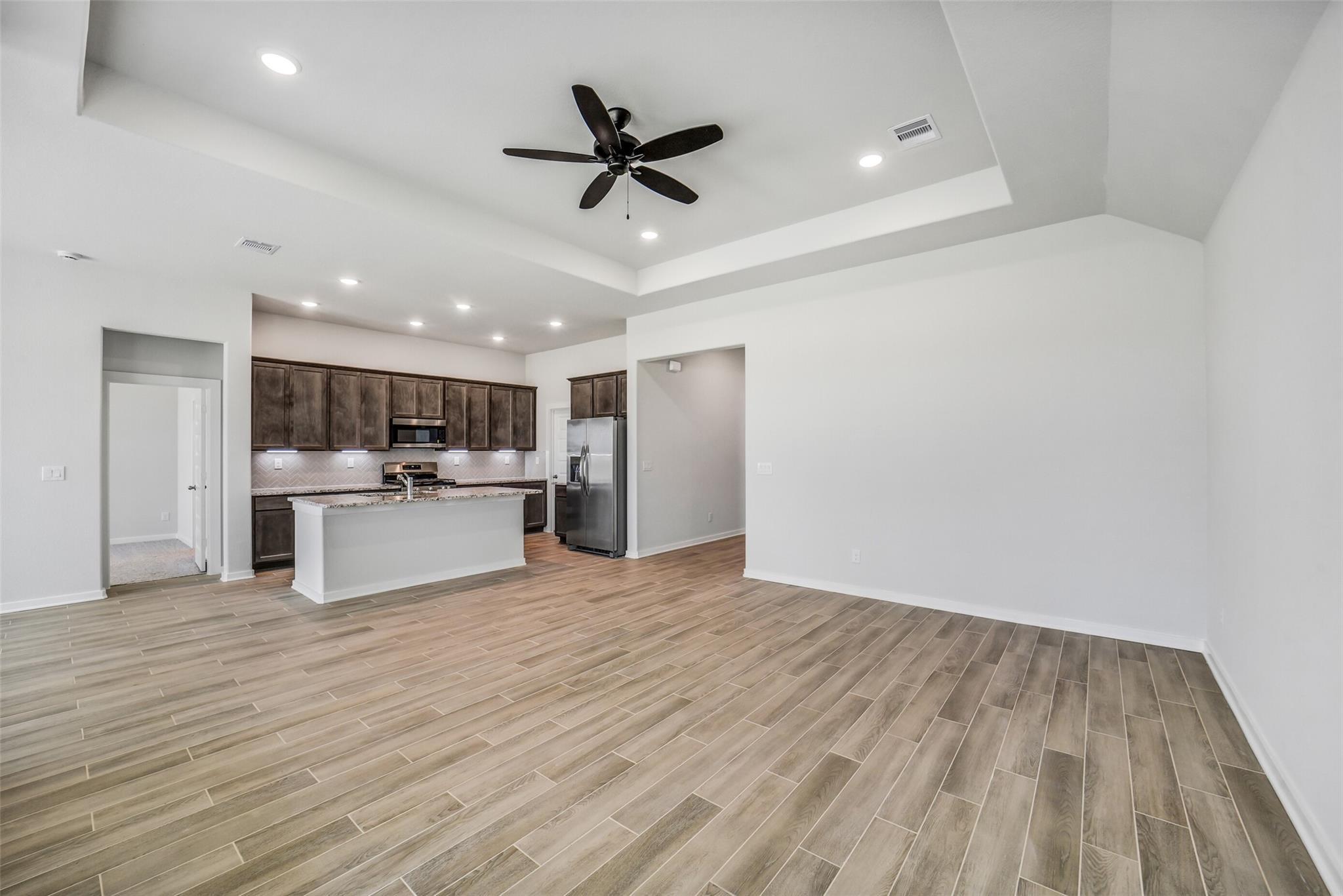 Open-concept kitchen with dark wood cabinets, white island, stainless fridge in living area with wood-grain tile floors and ceiling fan, Acadia C home, Magnolia TX