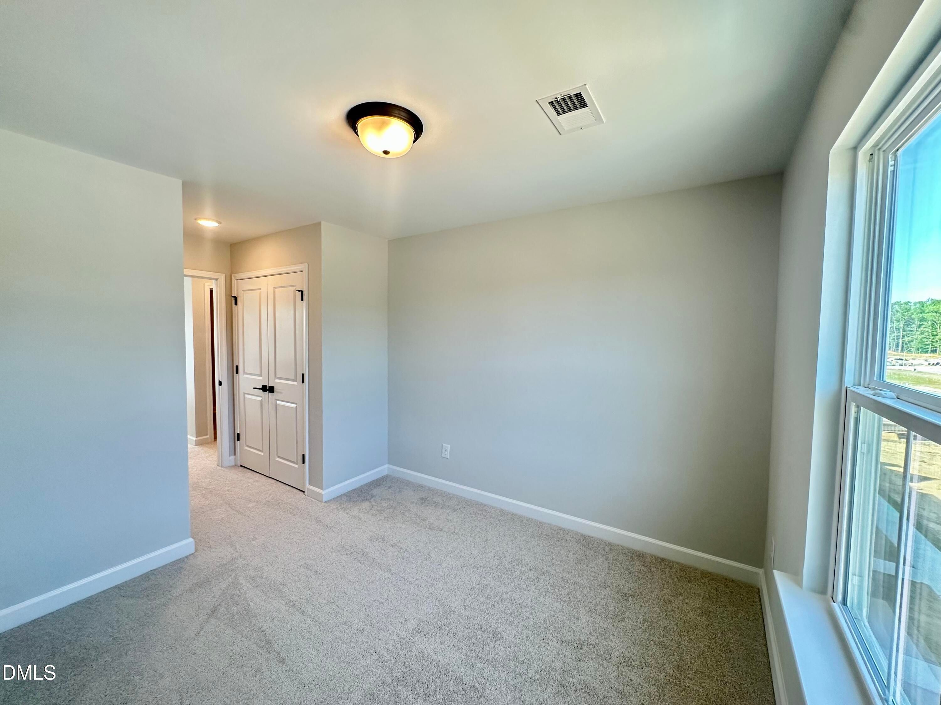 Spacious bedroom with light gray walls, beige carpet, double-door closet, and large window overlooking trees in Davidson Homes The Avery, Knightdale, NC