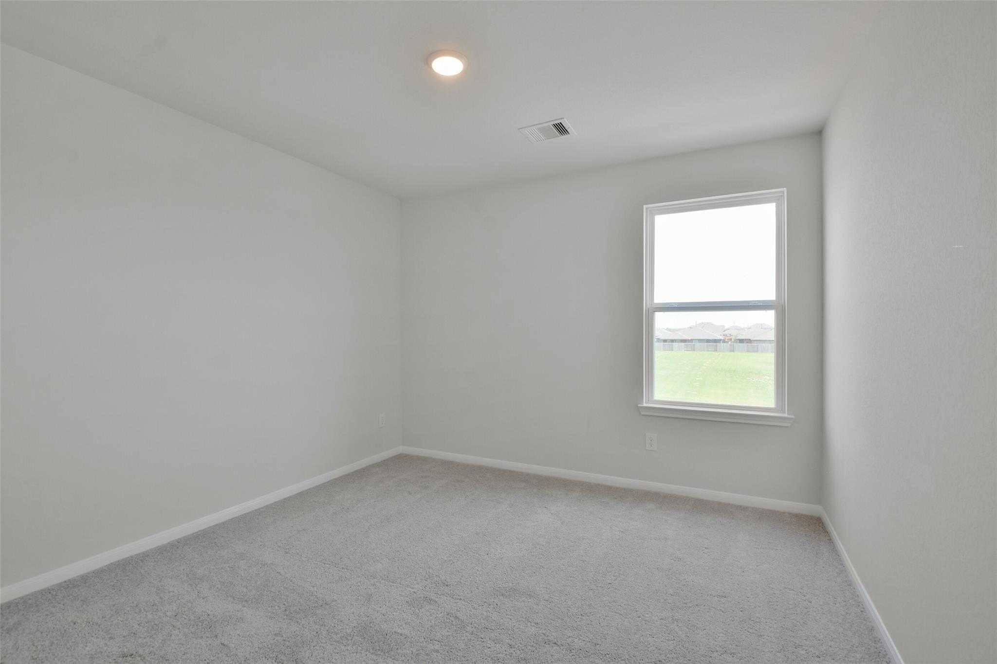 Bright empty bedroom with white walls, gray carpet, and window overlooking green yard in Davidson Homes The Tierra A, Dayton, Texas