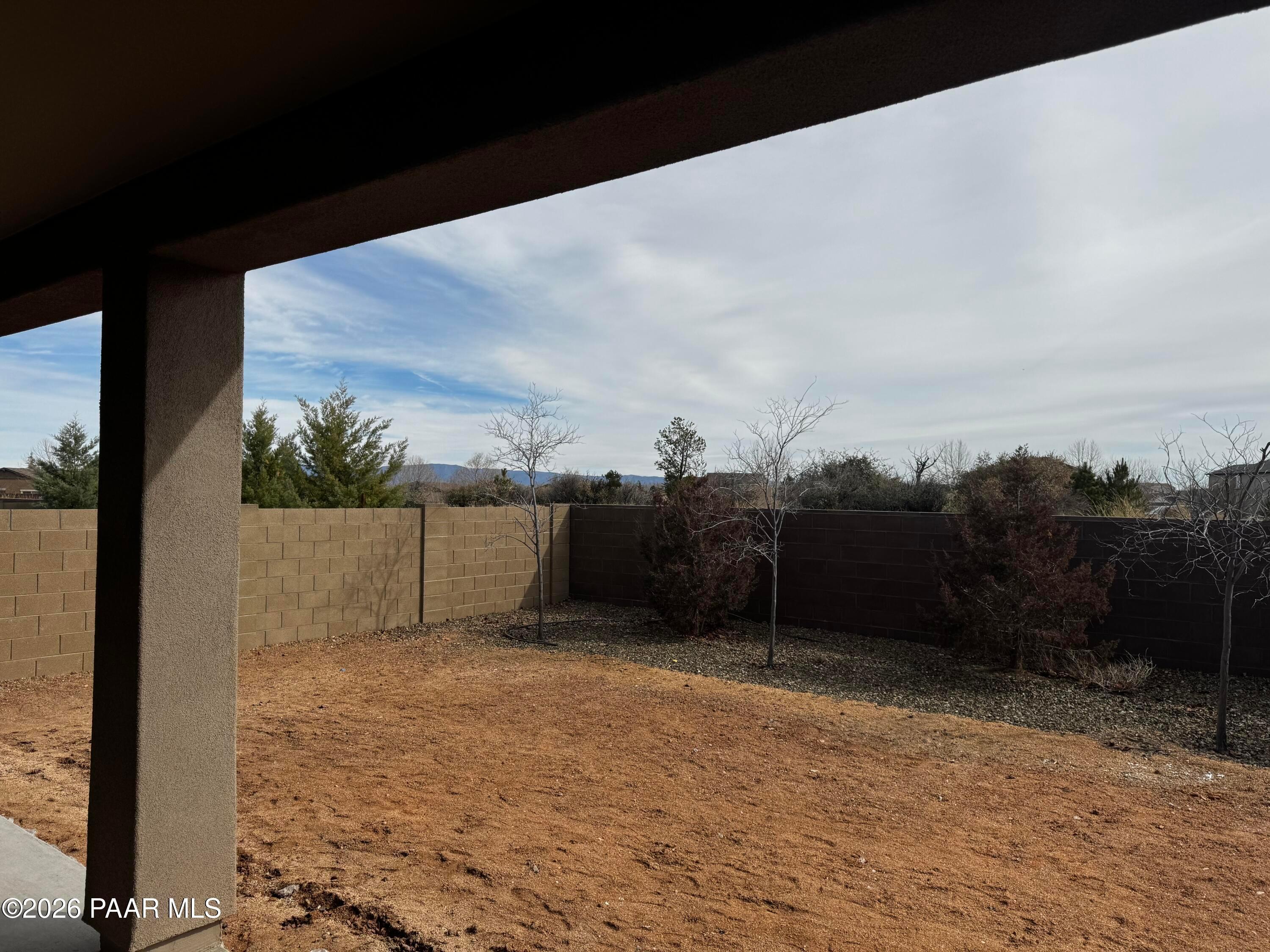Private backyard with tan block fence, young trees, shrubs, and mountain views under covered patio in Westwood, Prescott, Arizona