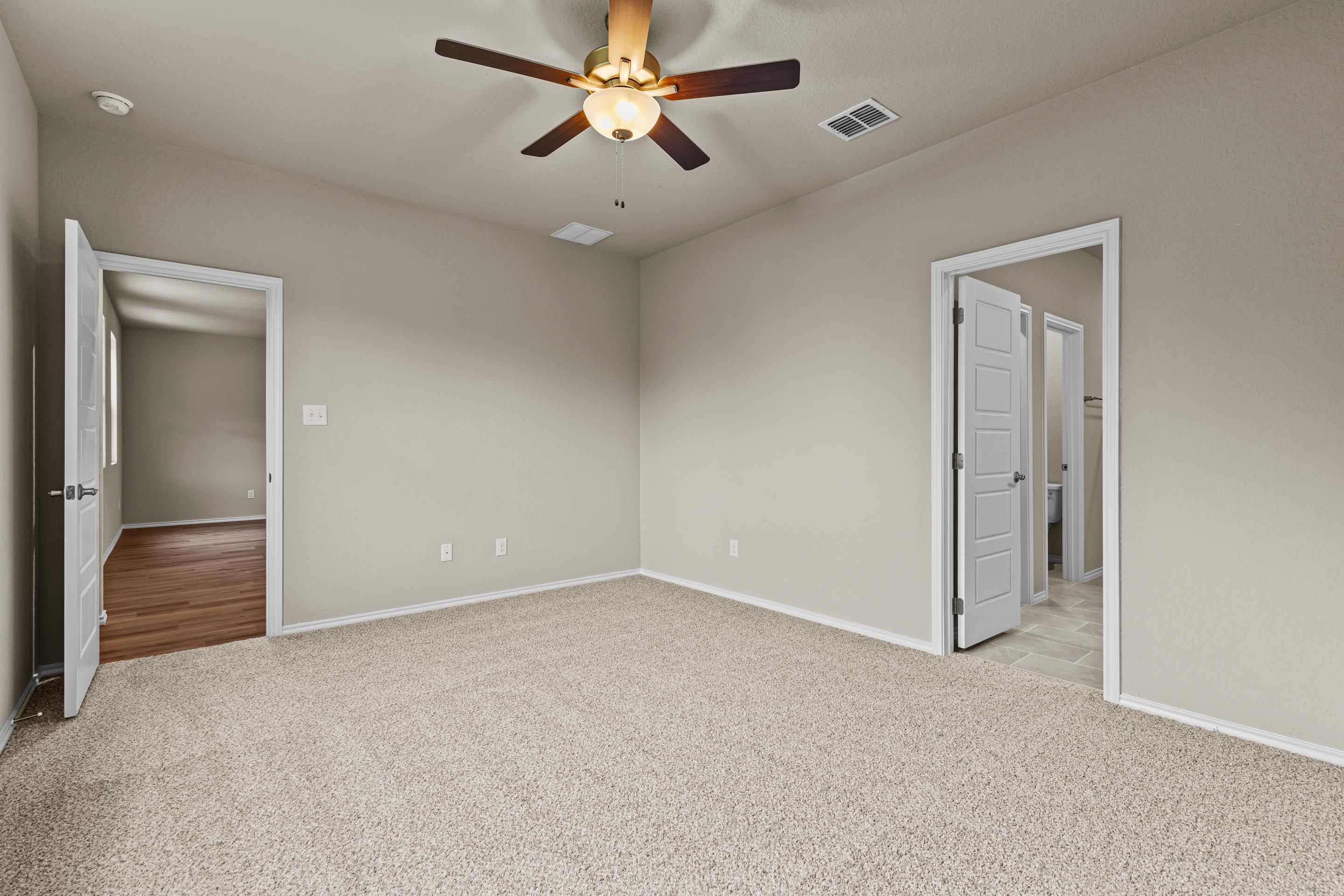 Spacious empty bedroom with beige carpet, ceiling fan, and doors to bathroom and hallway in Davidson Homes The Douglas F, San Antonio