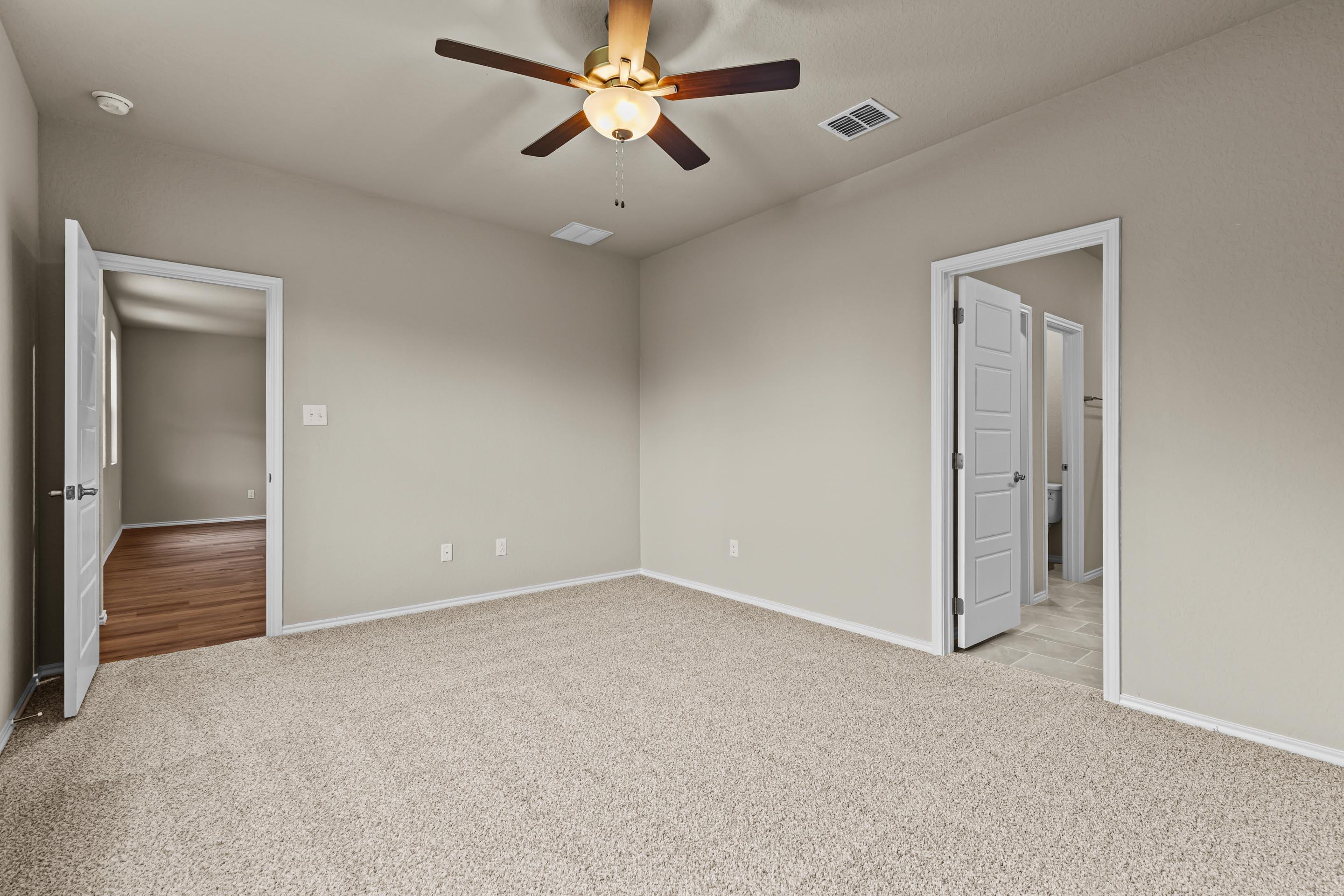 Spacious empty bedroom with beige carpet, ceiling fan, and doors to bathroom and hallway in Davidson Homes The Douglas F, San Antonio