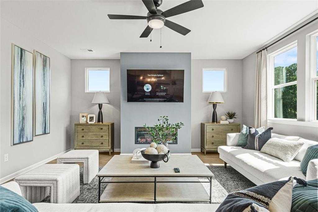 Cozy living room with white sectional sofa, mounted TV above fireplace, ceiling fan, and large windows in Davidson Homes The Hickory B, Hoschton, GA