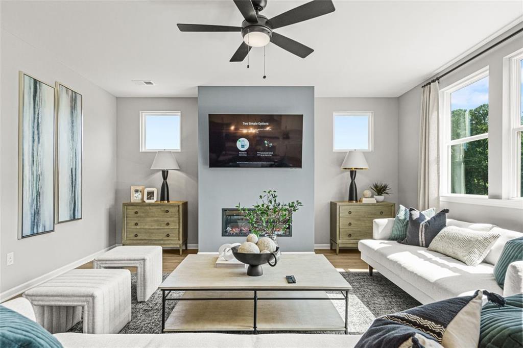 Cozy living room with white sectional sofa, mounted TV above fireplace, ceiling fan, and large windows in Davidson Homes The Hickory B, Hoschton, GA