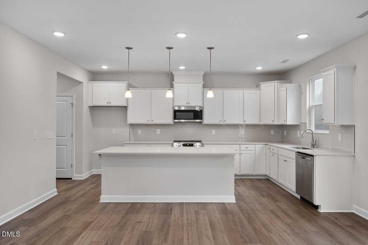 Modern white kitchen with large island, stainless steel appliances, pendant lights, and hardwood floors in The Hickory II A, Lillington, NC