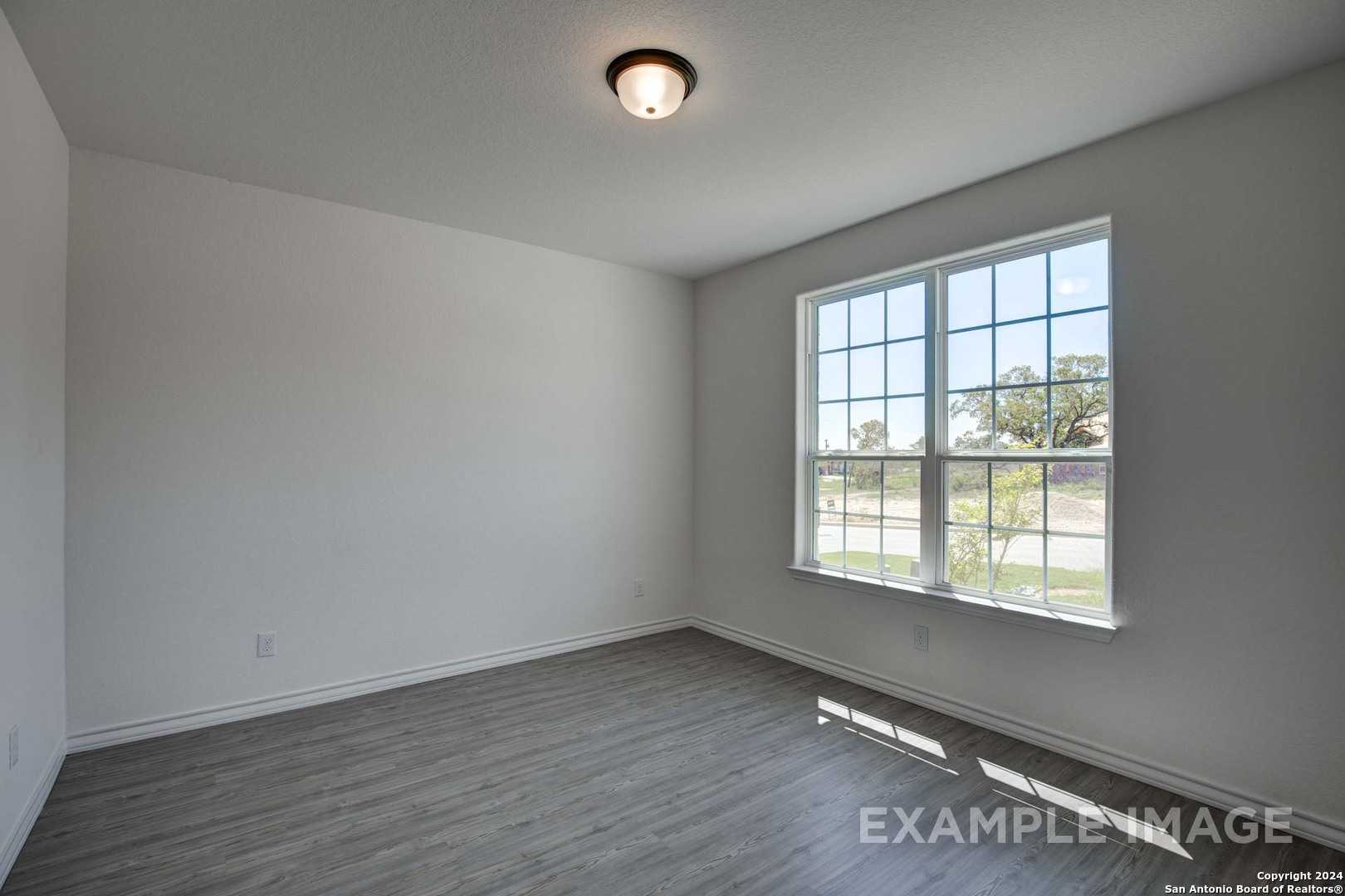 Bright empty bedroom with large grid window overlooking trees and gray laminate flooring in The Garner C, Castroville, Texas