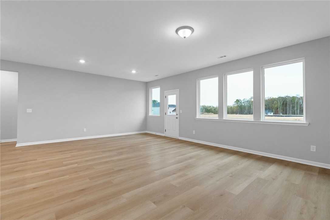Spacious living room with light hardwood floors, gray walls, and large windows overlooking trees in Davidson Homes The Luna B, Opelika, Alabama