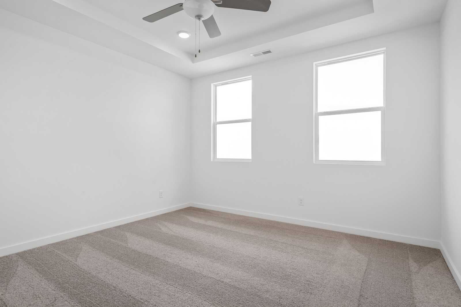 Spacious empty bedroom in The Asheville design with white walls, two large windows, ceiling fan, and neutral carpet flooring