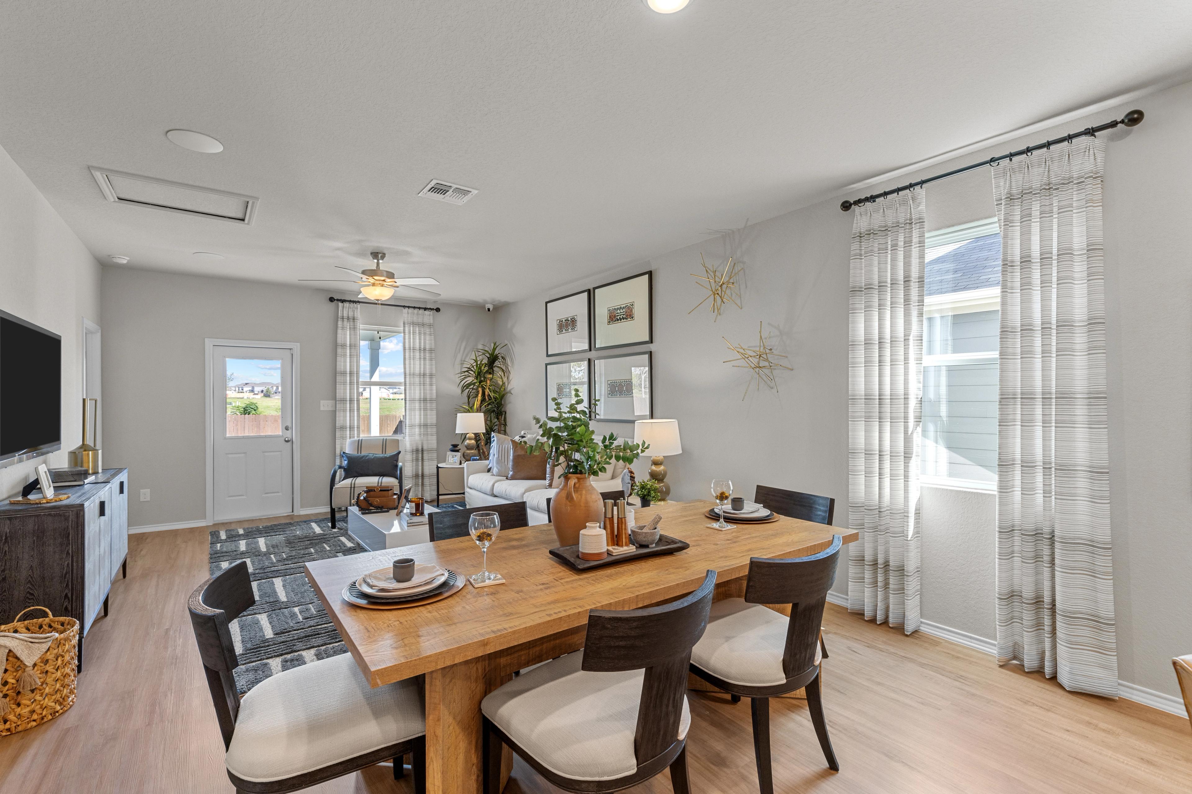 Inviting dining area in The Sabine B home design featuring rustic wooden table, upholstered chairs, sheer curtains, and TV wall