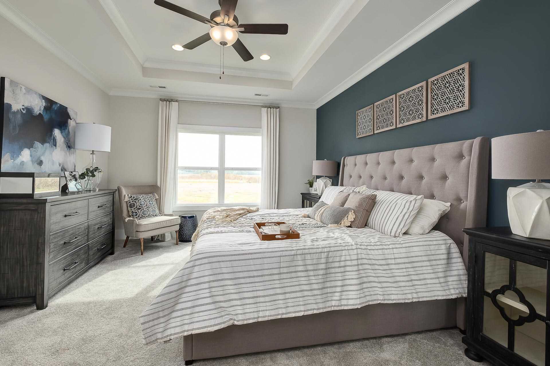 Spacious master bedroom in The Everett C with tufted upholstered bed, navy accent wall, ceiling fan, and large window