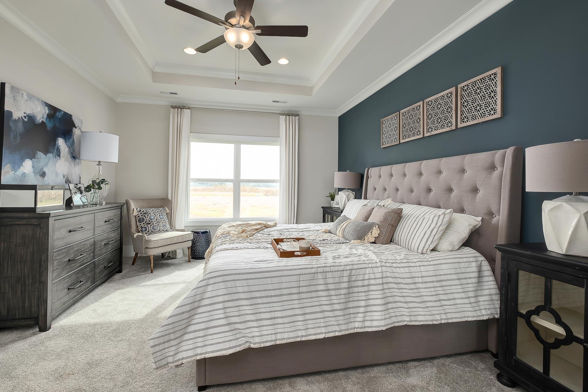 Spacious master bedroom in The Everett featuring tufted upholstered bed, navy accent wall, ceiling fan, armchair, and window view