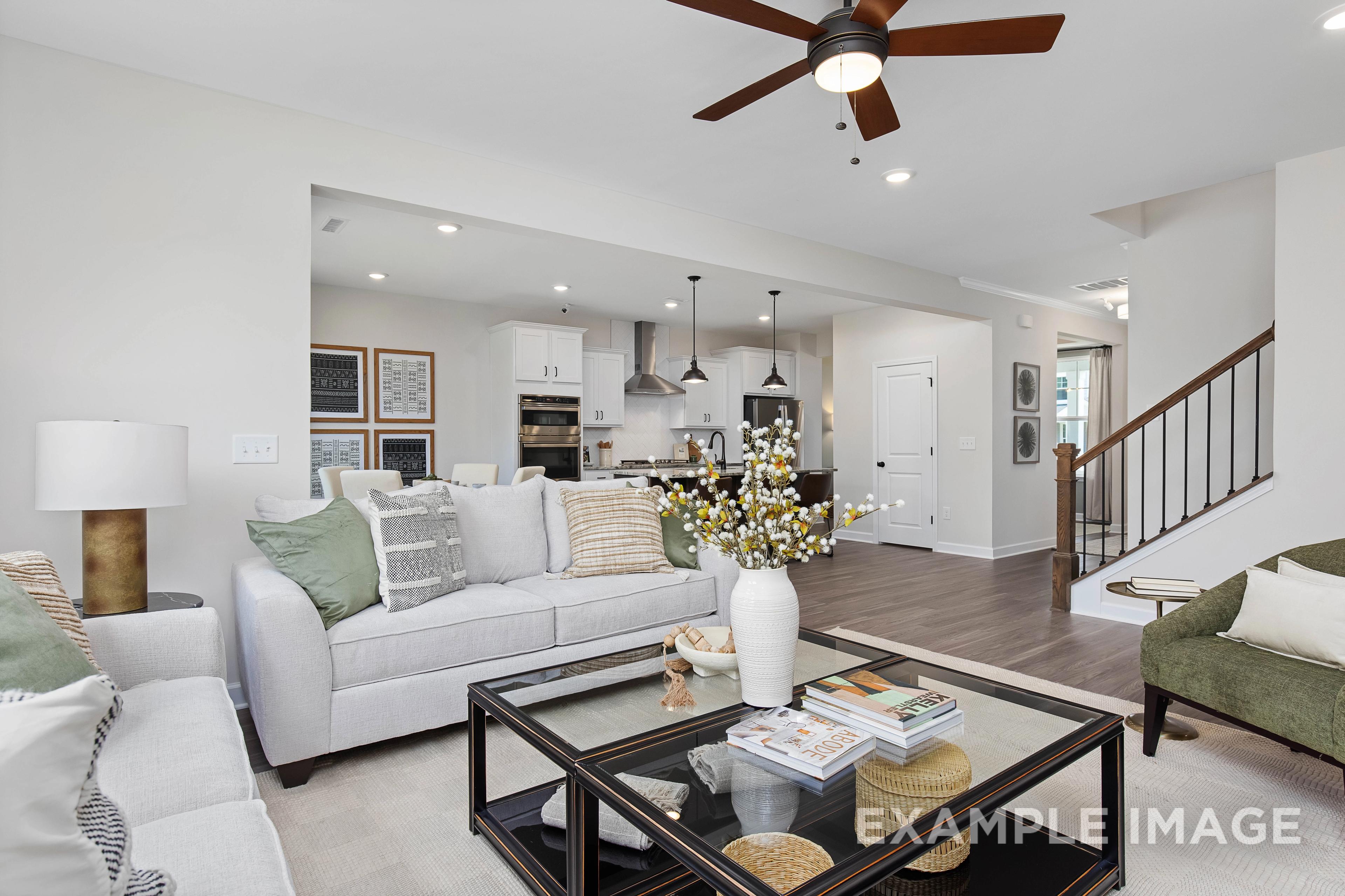 Spacious open-concept living room in The Willow B home with gray sectional sofas, glass coffee table, potted flowers, and adjacent modern kitchen