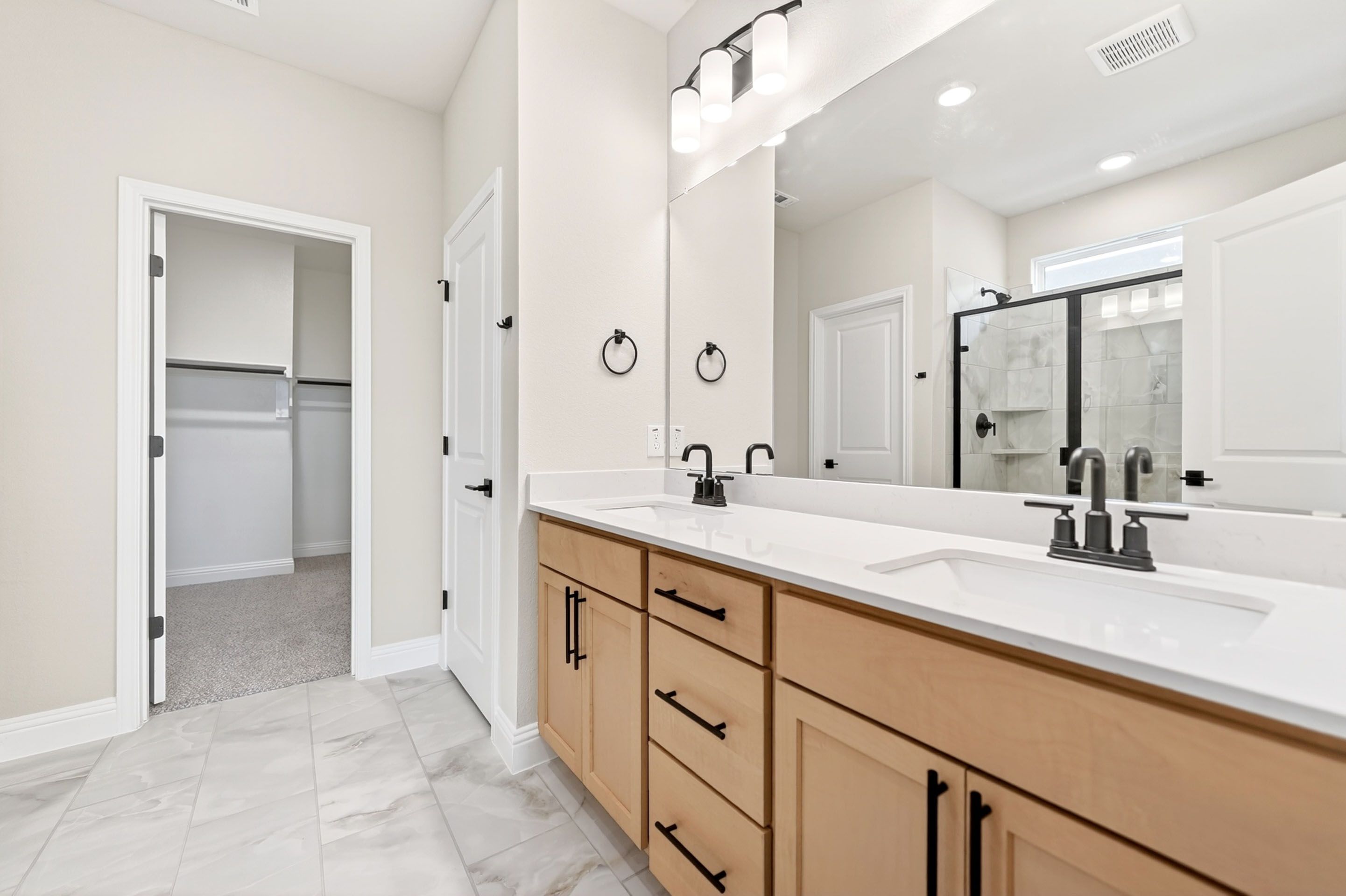 Modern master bathroom in The Holly home design featuring dual vanities, light wood cabinets, and a walk-in closet.