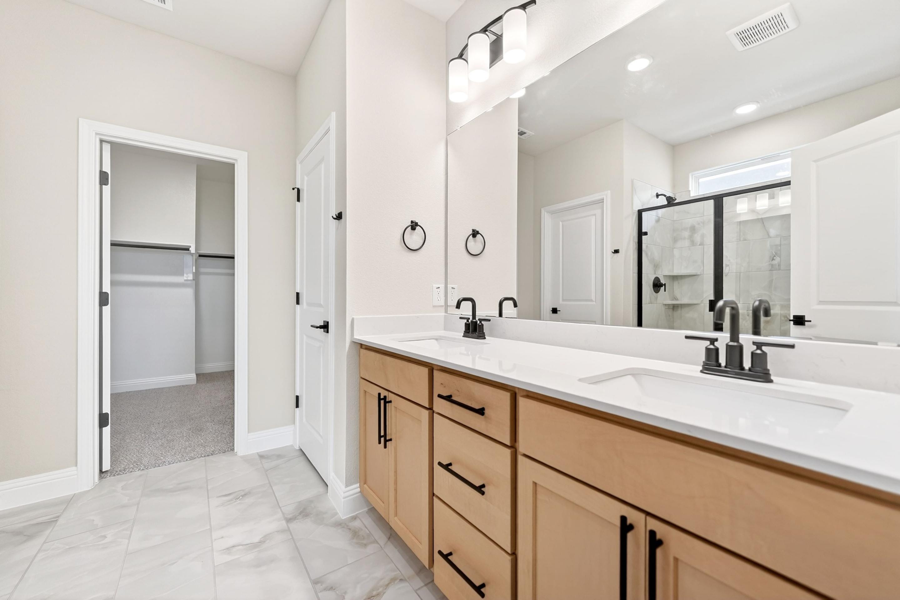 Modern master bathroom in The Holly home design featuring dual vanities, light wood cabinets, and a walk-in closet.