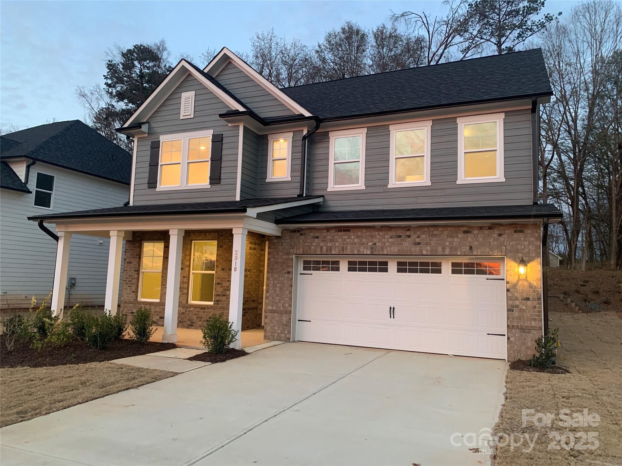 Twilight view of 2-story gray-sided 4-bedroom home with 2-car garage, lit windows, and front porch in Enclave at Belmont, NC
