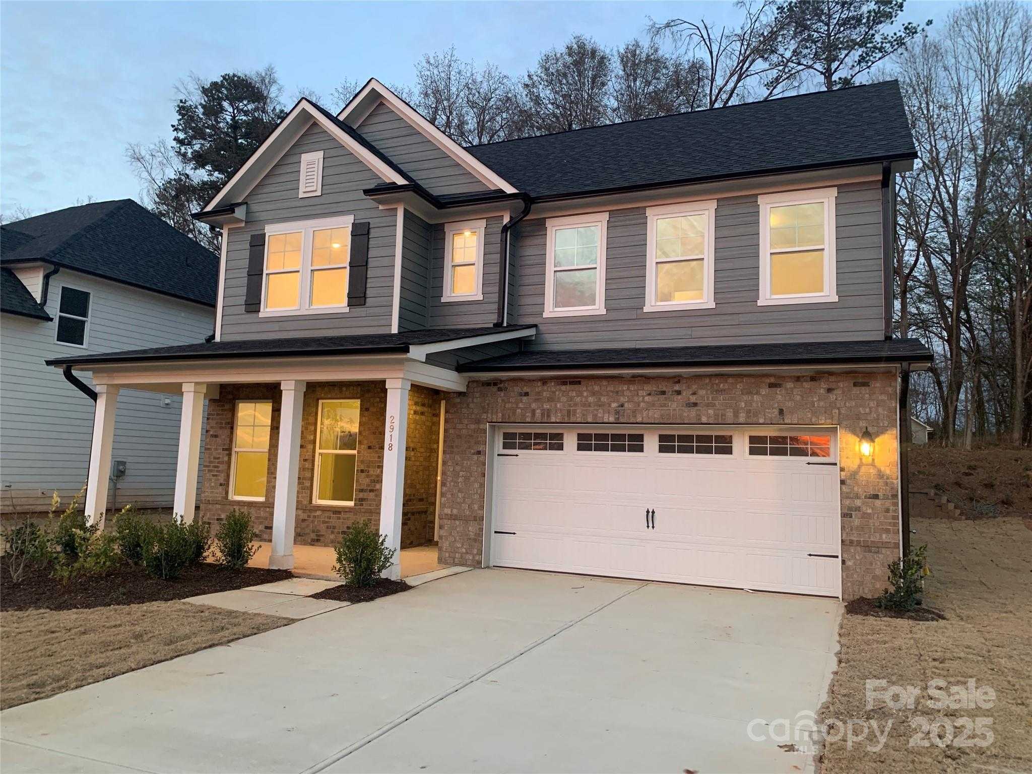 Twilight view of 2-story gray-sided 4-bedroom home with 2-car garage, lit windows, and front porch in Enclave at Belmont, NC