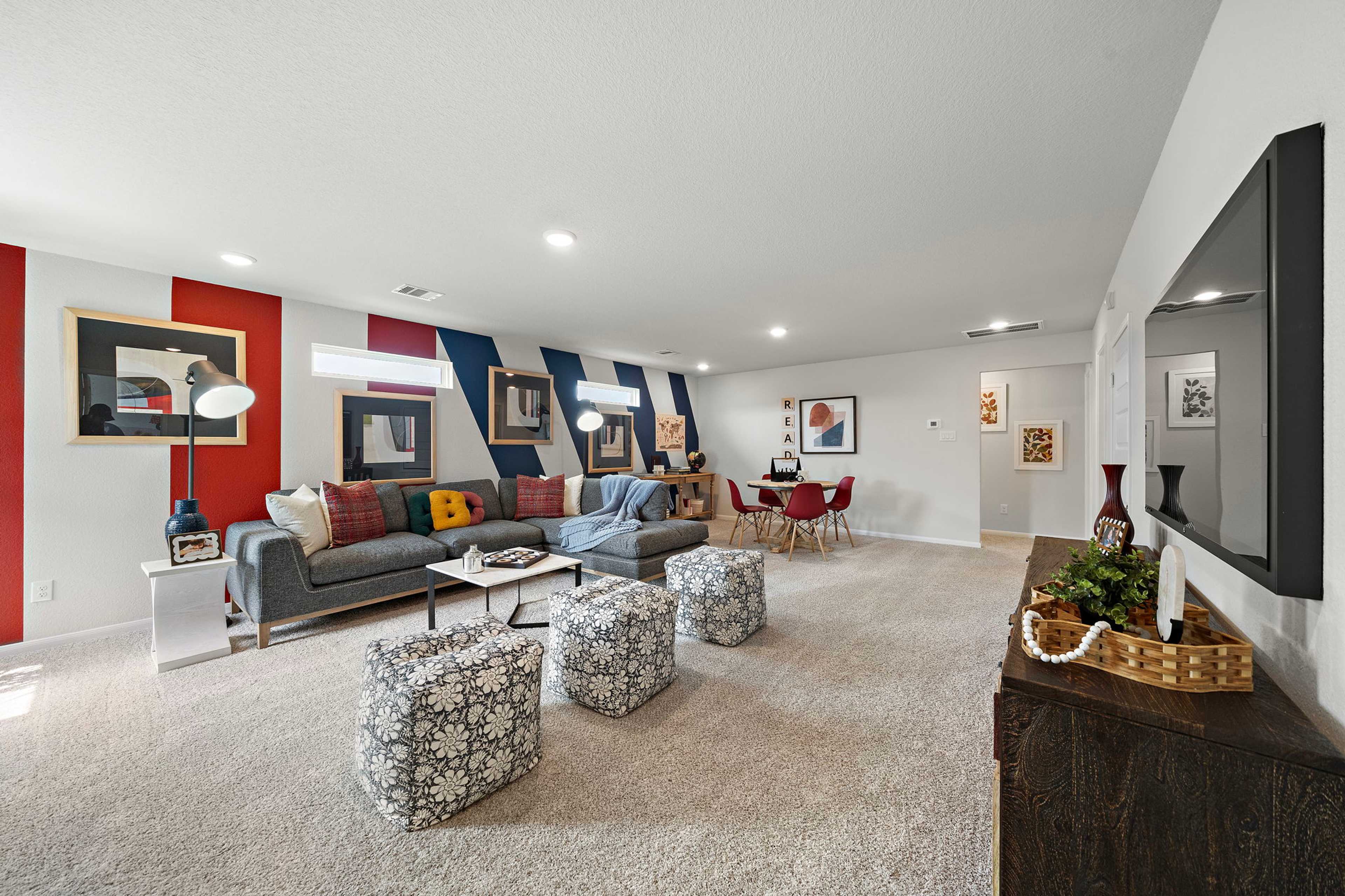 Spacious living room in Liberty Estates home, Cleveland Texas, with gray sectional sofa, colorful abstract wall art, red dining chairs, and mounted TV