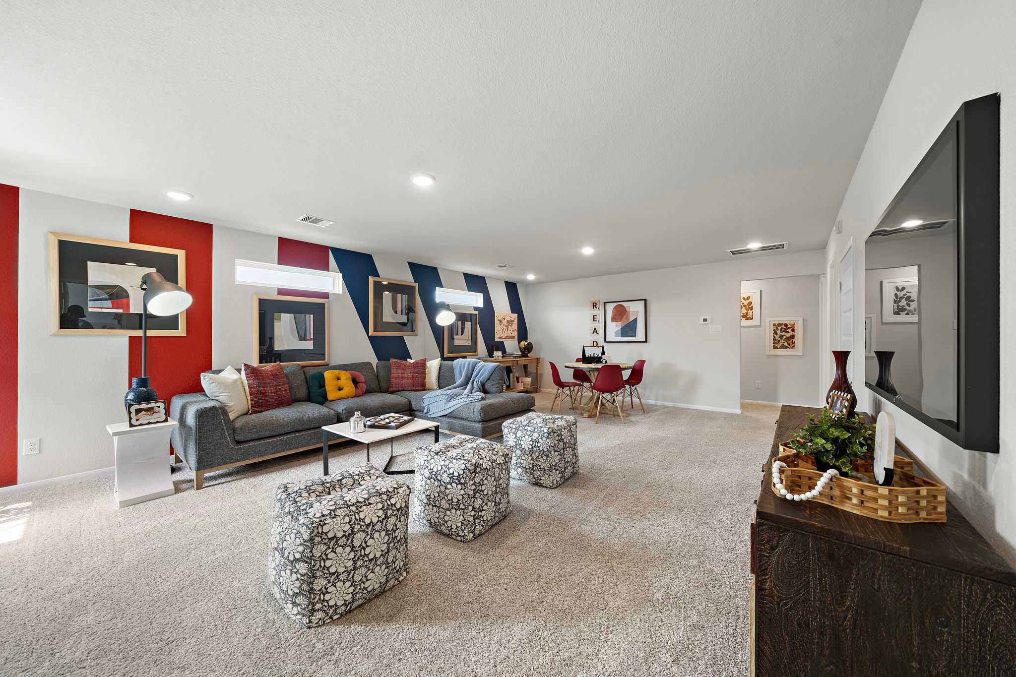 Spacious living room in Liberty Estates home, Cleveland Texas, with gray sectional sofa, colorful abstract wall art, red dining chairs, and mounted TV