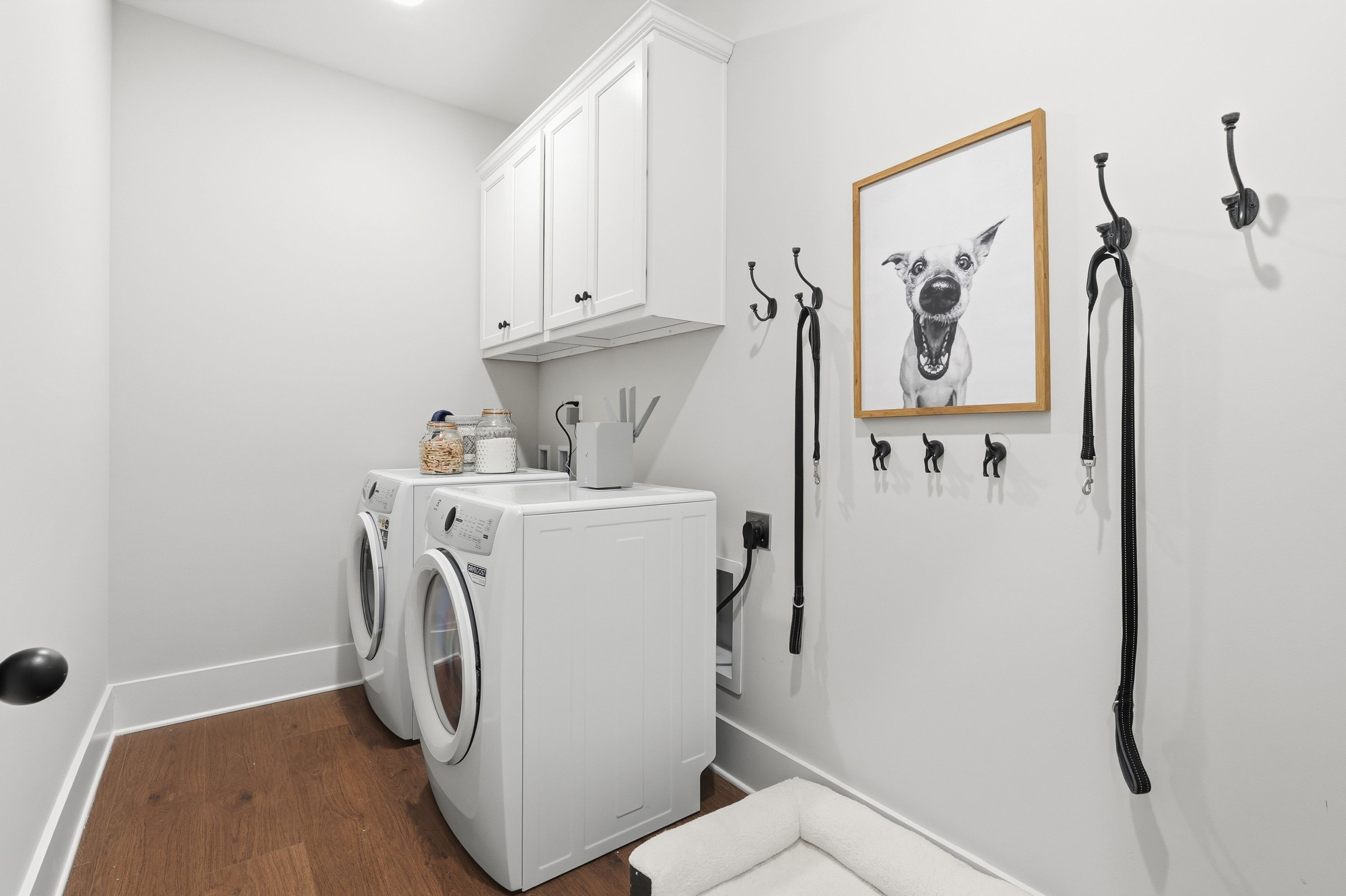 Modern laundry room at Noble Ridge in Cullman Alabama with white washer dryer, cabinets, wall hooks, dog portrait, and pet bed on hardwood floors