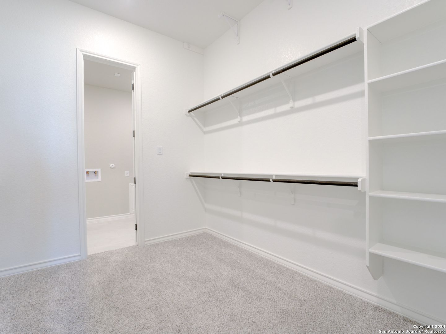 Spacious walk-in closet with built-in white shelves and hanging rods next to bathroom in Davidson Homes The Summerlin A, Castroville, Texas