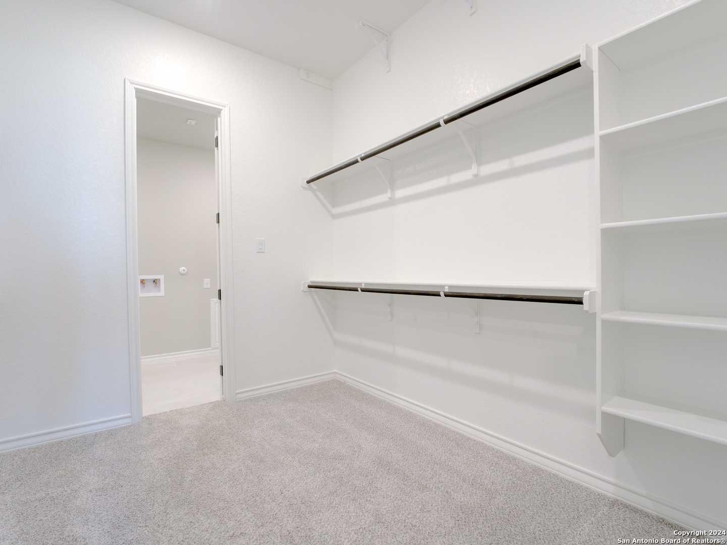 Spacious walk-in closet with built-in white shelves and hanging rods next to bathroom in Davidson Homes The Summerlin A, Castroville, Texas