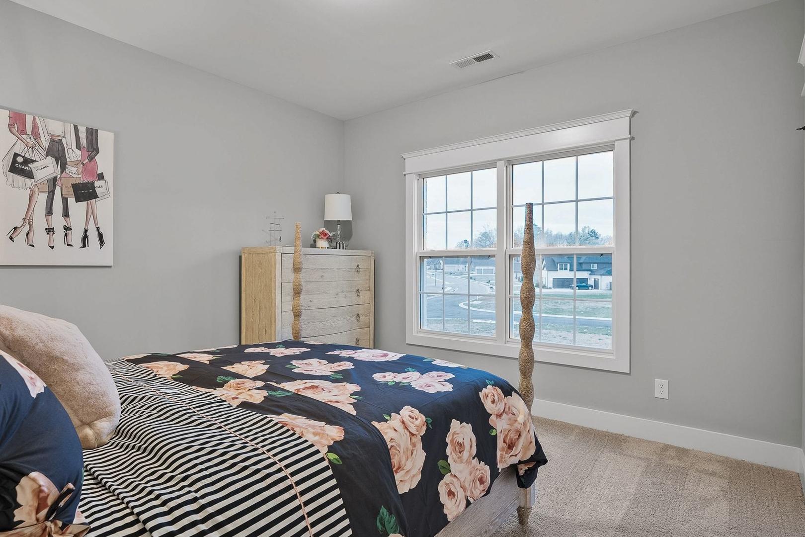 Spacious bedroom in The Oxford with floral bedding on king bed, gray walls, dresser, window view, and fashion artwork