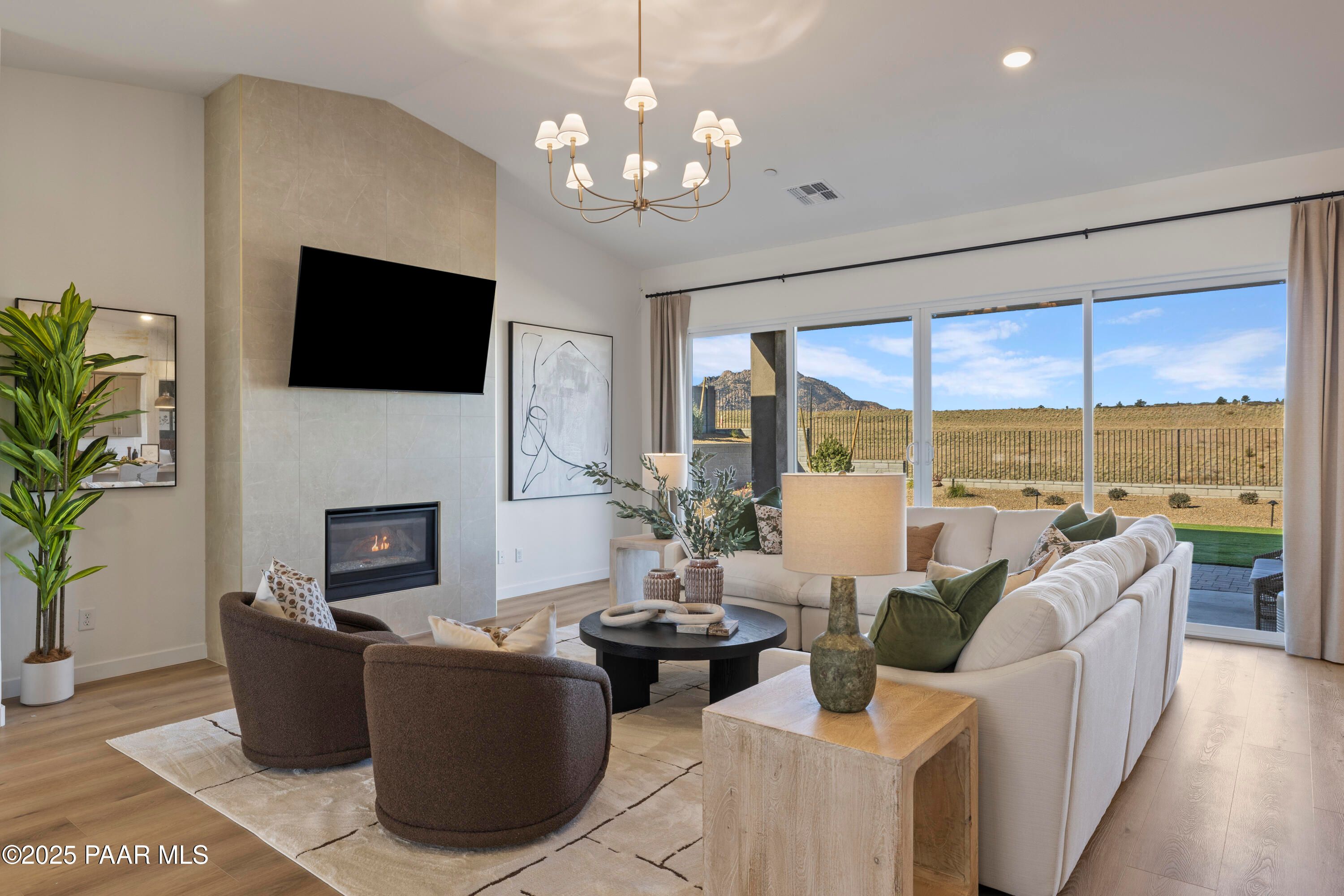 Cozy living room with stone gas fireplace, mounted TV, chandelier, and desert views via sliding glass doors in Davidson Homes The Blaze D, Prescott, Arizona