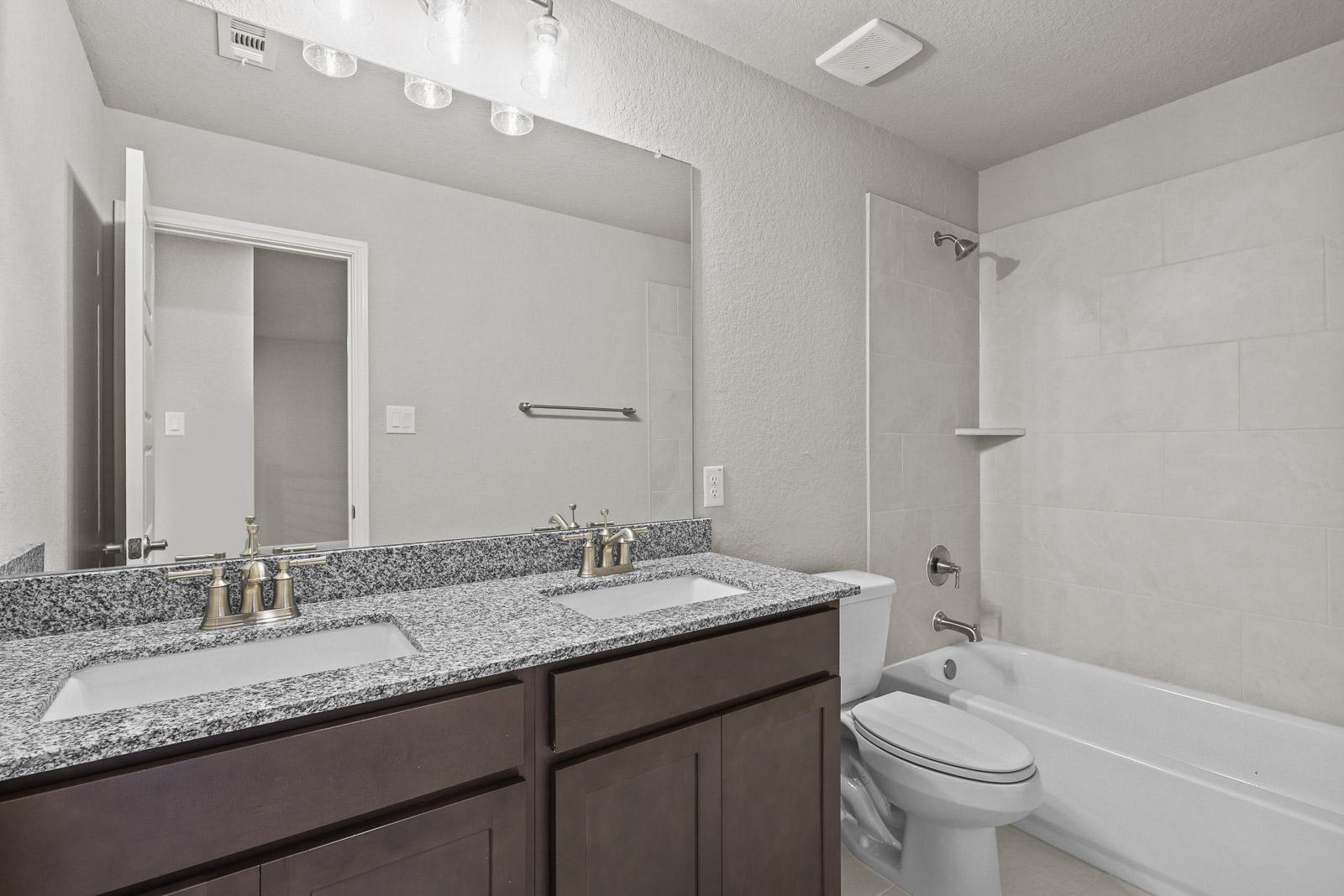 Spacious bathroom in The Jennings featuring a dual-sink granite vanity, dark wood cabinets, and a tiled shower-tub combo.