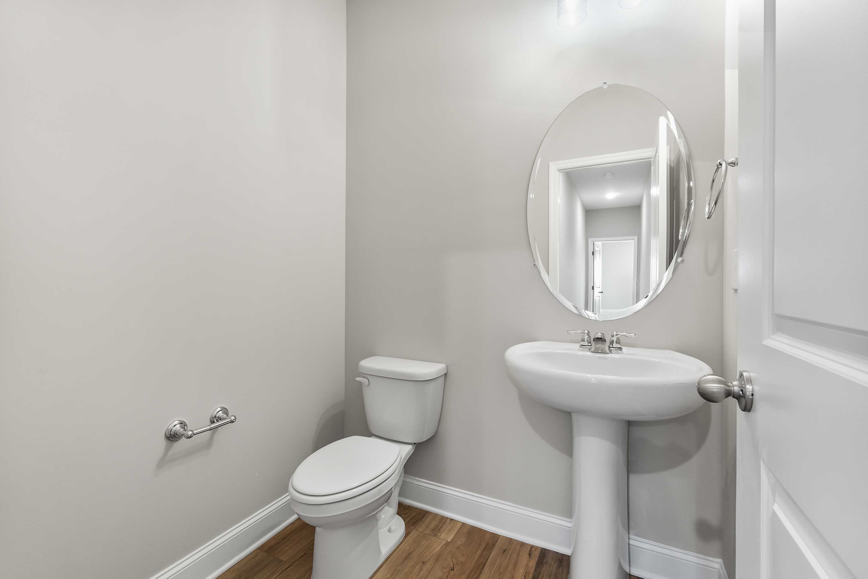 Compact powder room in The Valencia featuring pedestal sink, round mirror, white toilet, neutral walls, and hardwood floors