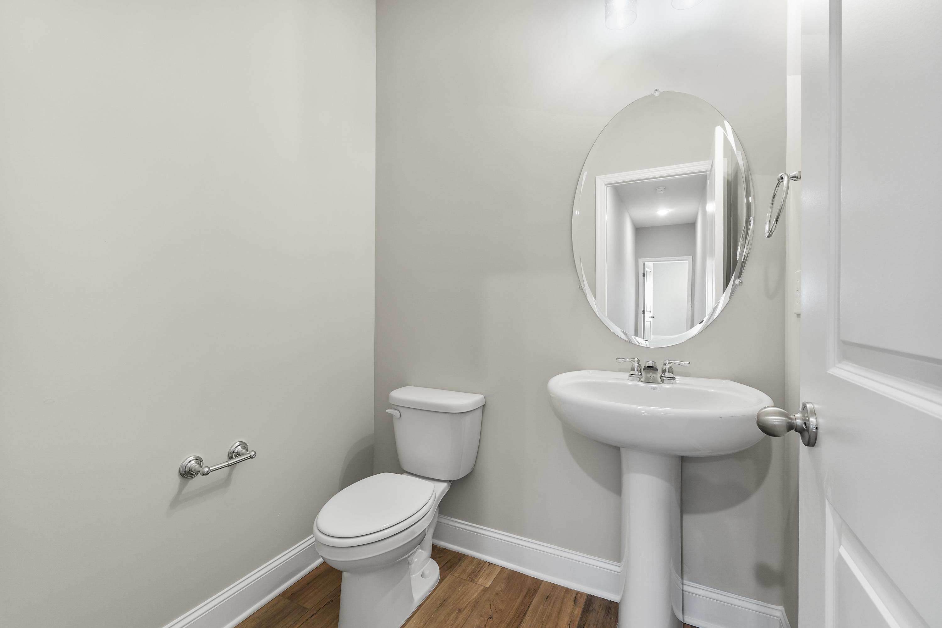 Compact powder room in The Valencia featuring pedestal sink, round mirror, white toilet, neutral walls, and hardwood floors