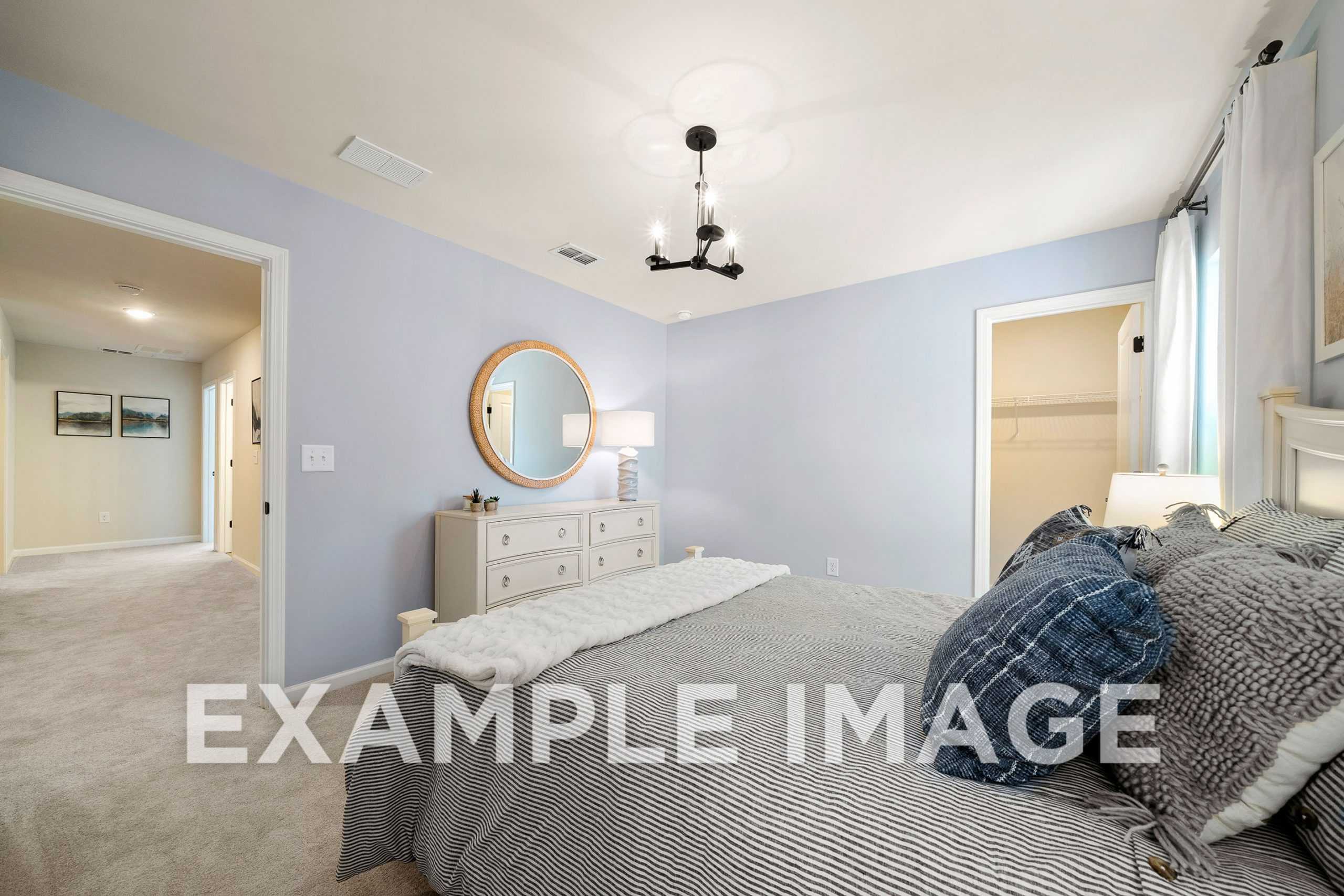 Spacious master bedroom in The Hickory A with light blue walls, gray bedding, white dresser, round mirror, and chandelier lighting