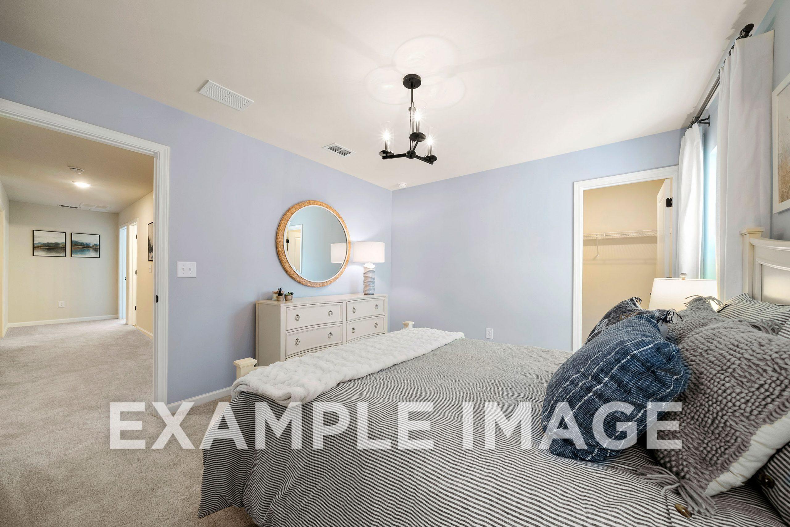 Spacious master bedroom in The Hickory B with light blue walls, king bed in gray and blue bedding, white dresser, round mirror, and chandelier