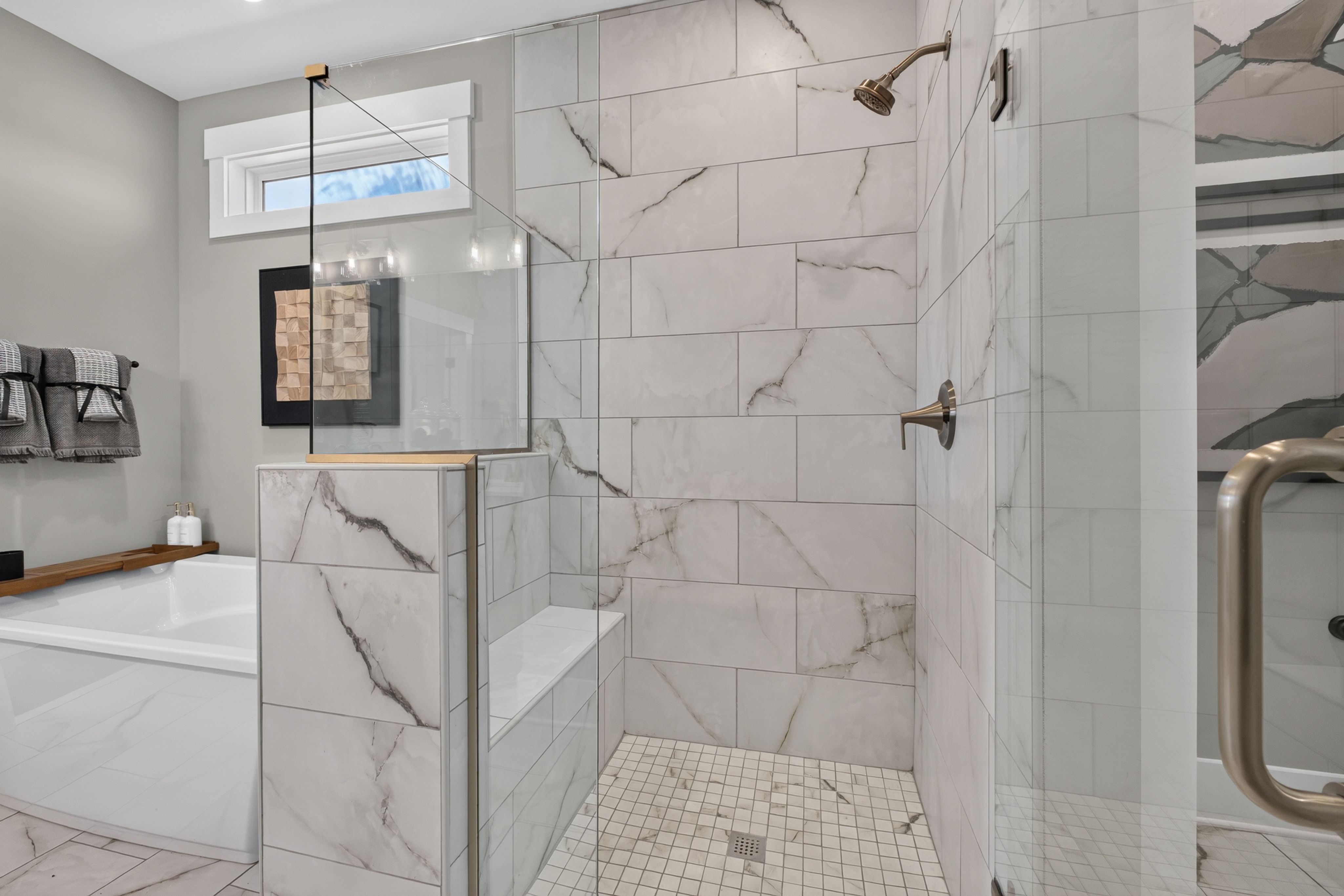 Marble-tiled walk-in shower with glass panel and bench beside soaking tub in Evergreen Mill master bath by Davidson Homes, Madison AL