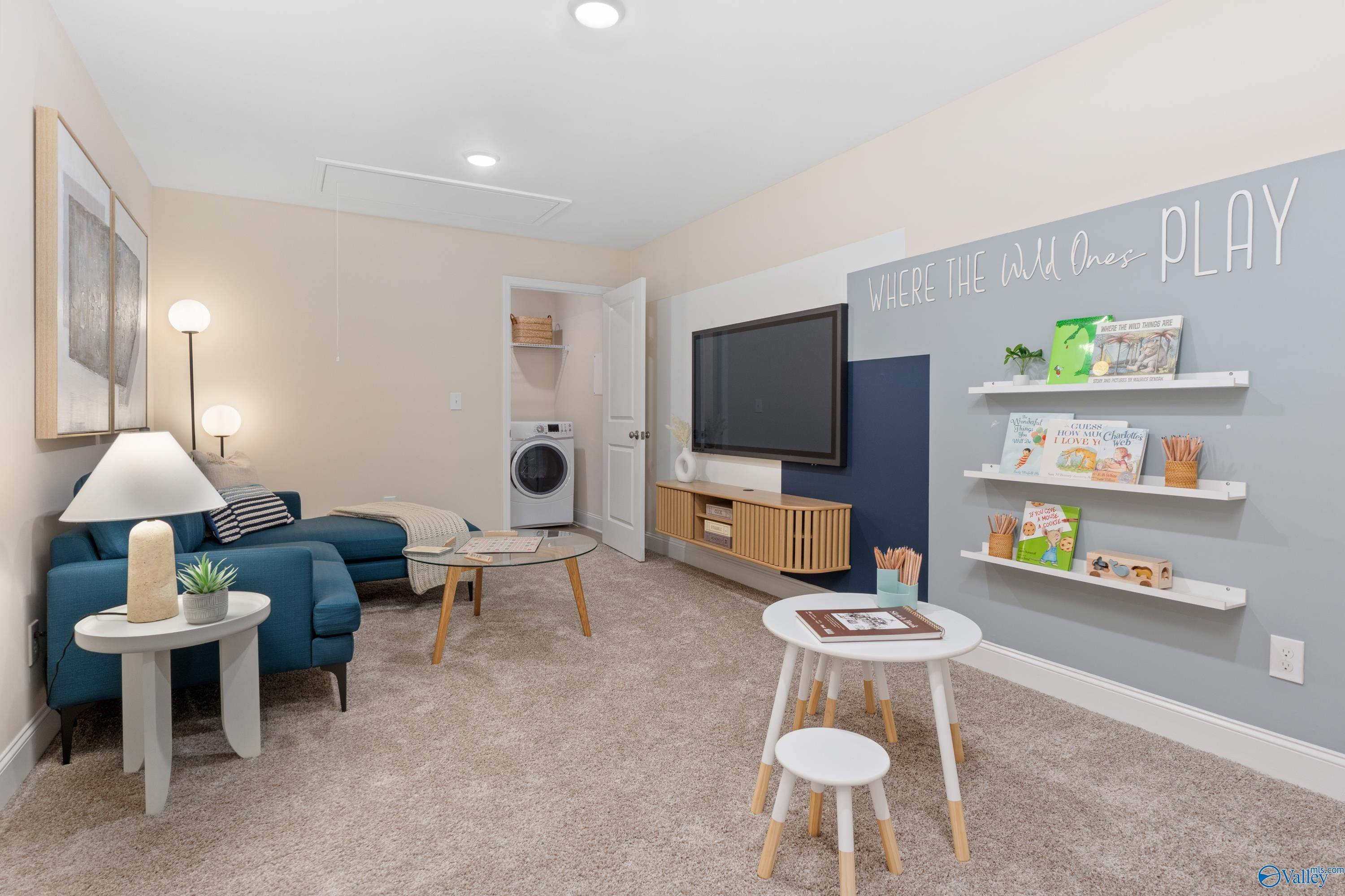 Cozy basement playroom with navy sectional sofa, mounted TV, children's bookshelves, and laundry area in Davidson Homes The Camden, Huntsville AL