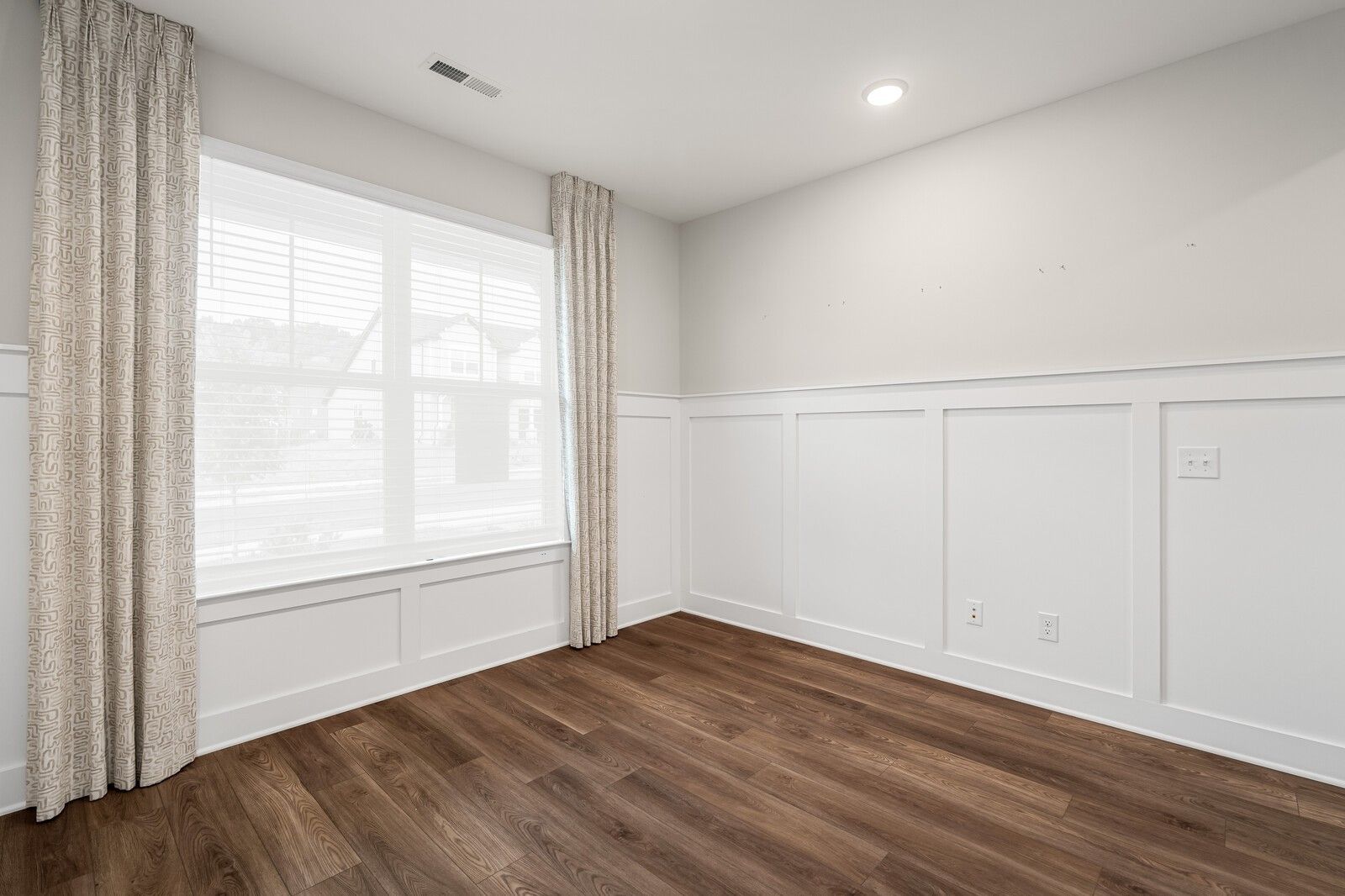 Bright bedroom corner with white wainscot walls, large window with beige curtains, and hardwood floors in Davidson Homes The Ridgeport, Gallatin TN