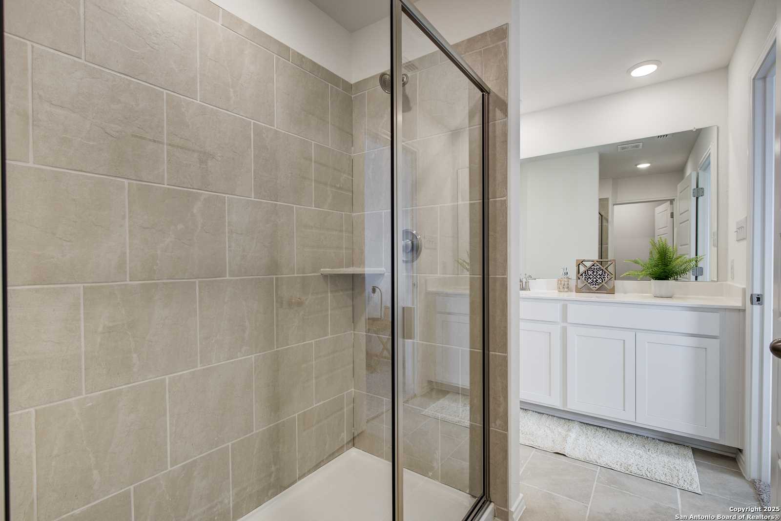 Modern master bathroom with frameless glass shower, beige tile walls, and white vanity in Davidson Homes The Trinity A, San Antonio