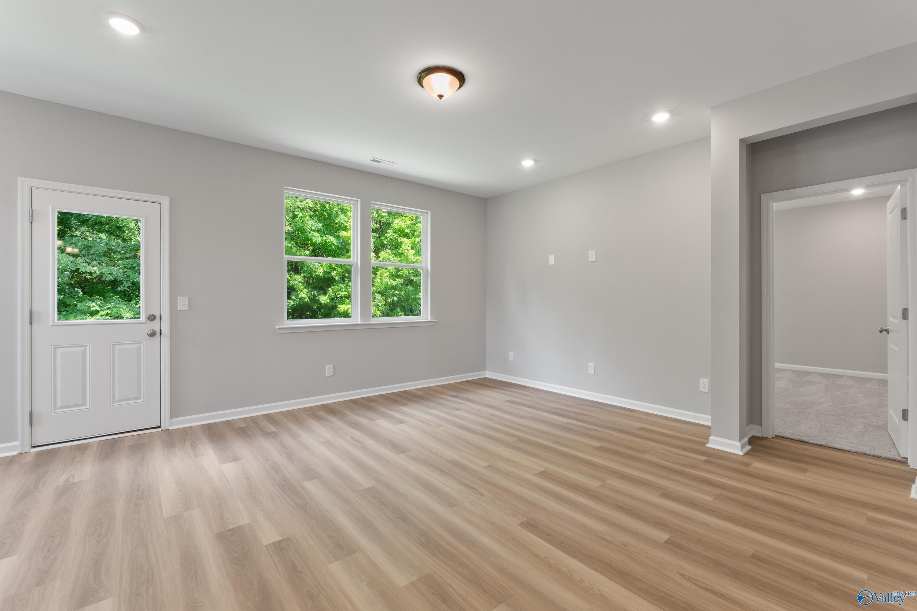 Bright empty living room with gray walls, luxury vinyl plank flooring, and windows overlooking green trees in Davidson Homes The Phoenix, Hazel Green, Alabama