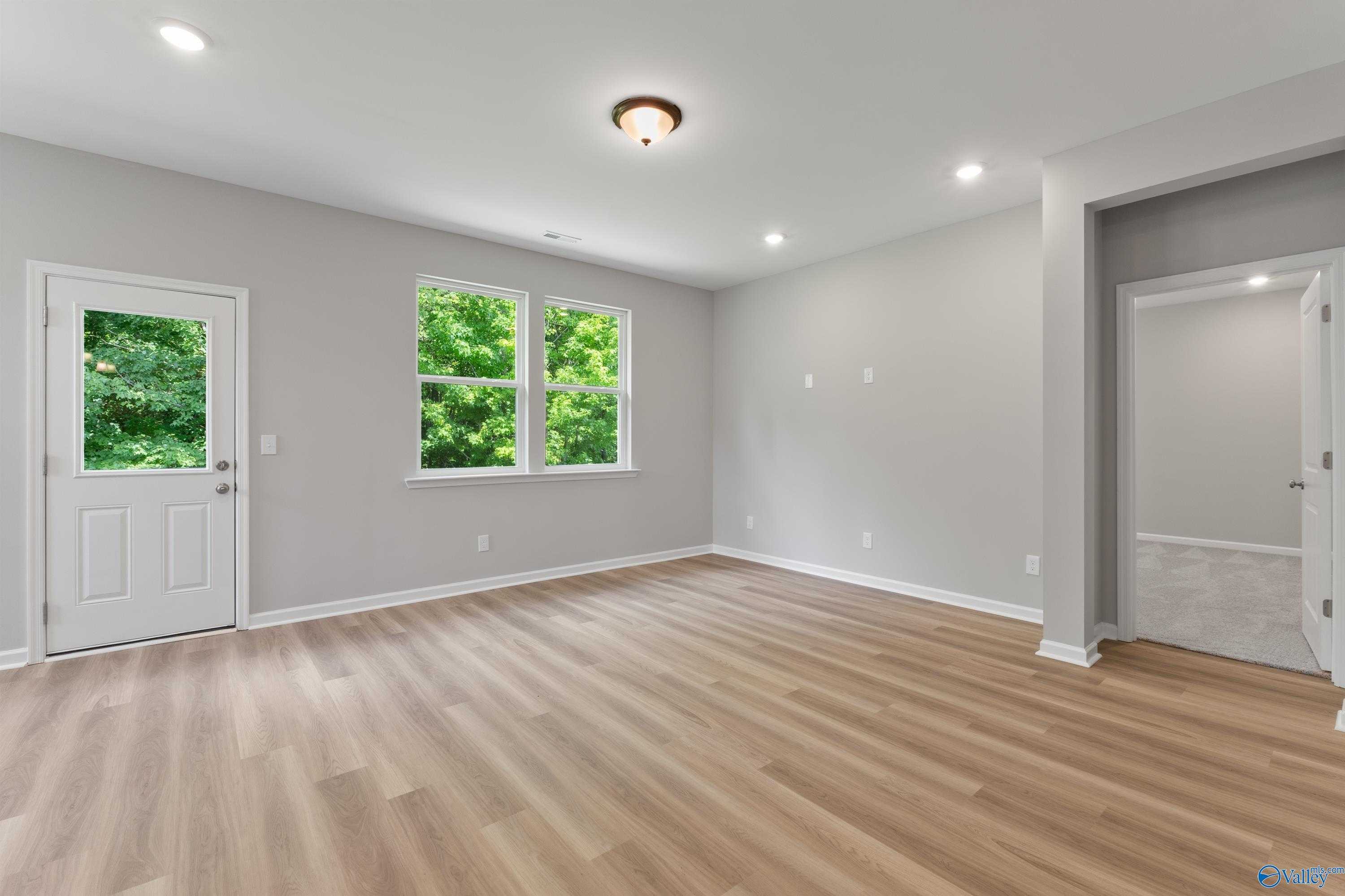 Spacious empty room with gray walls, light wood floors, large windows to green trees, and white doors in Davidson Homes The Phoenix, Hazel Green AL