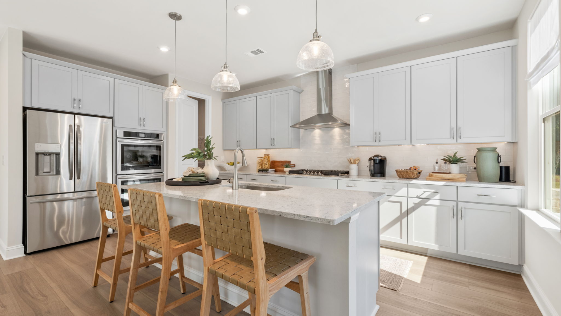 Modern white kitchen island with woven bar stools, stainless appliances, and pendant lights in Davidson Homes Fern Hollow model home