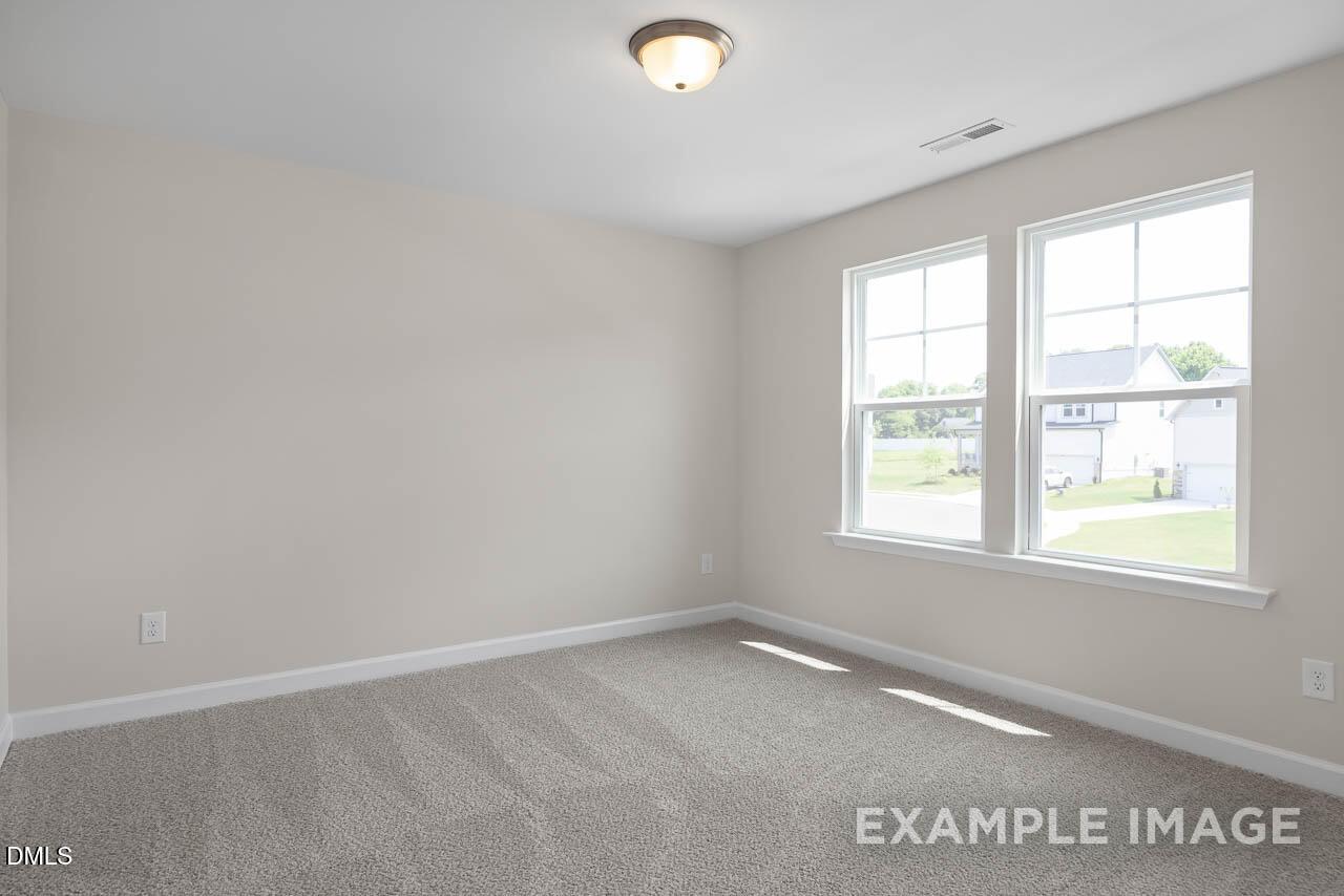 Bright empty bedroom with beige walls, neutral carpet, and large windows overlooking backyard in Davidson Homes The Willow D, Lillington, NC