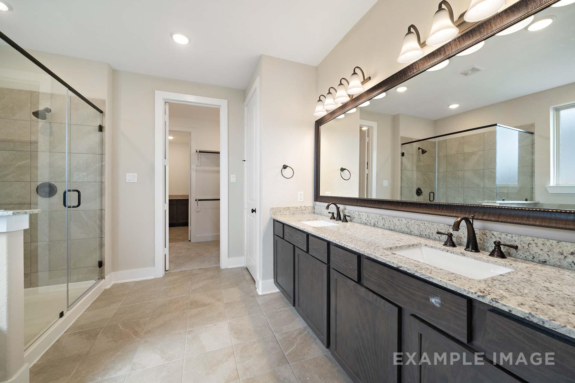 Spacious master bathroom in The Elizabeth A with frameless glass shower, double granite vanity, dark cabinets, and large lighted mirror