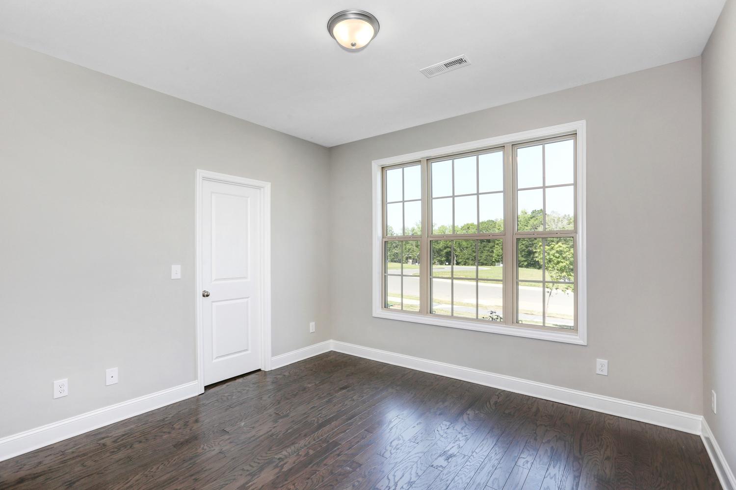 Spacious master bedroom in The Montgomery featuring light gray walls, dark hardwood floors, and large triple window with tree views