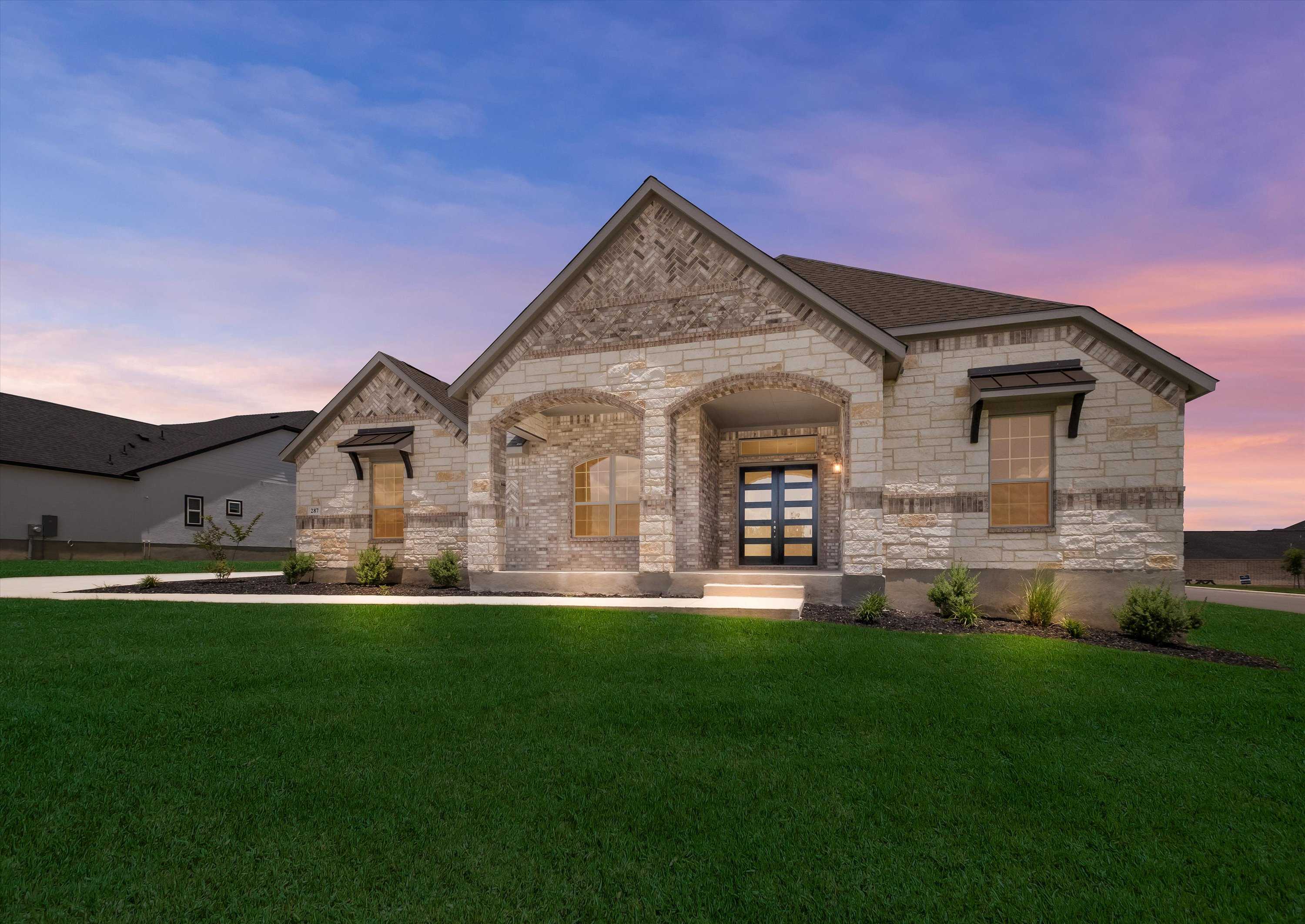 Modern stone facade single-story home with arched entry, double doors, and lush front yard at dusk in Potranco Oaks, Castroville, Texas
