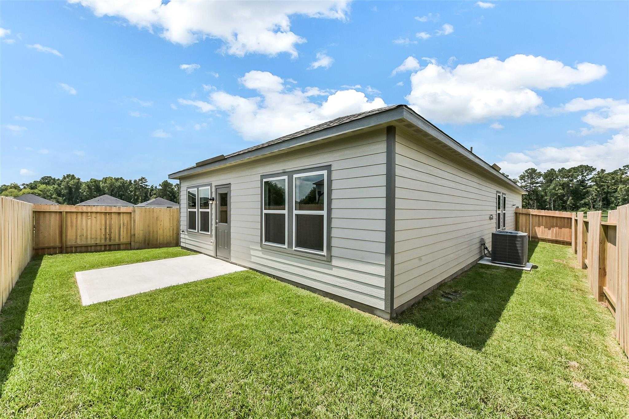 Modern single-story home with beige siding, fenced grassy backyard, concrete patio, and AC unit in The Villages at WestPointe, Dayton, Texas - Davidson Homes The Frio F